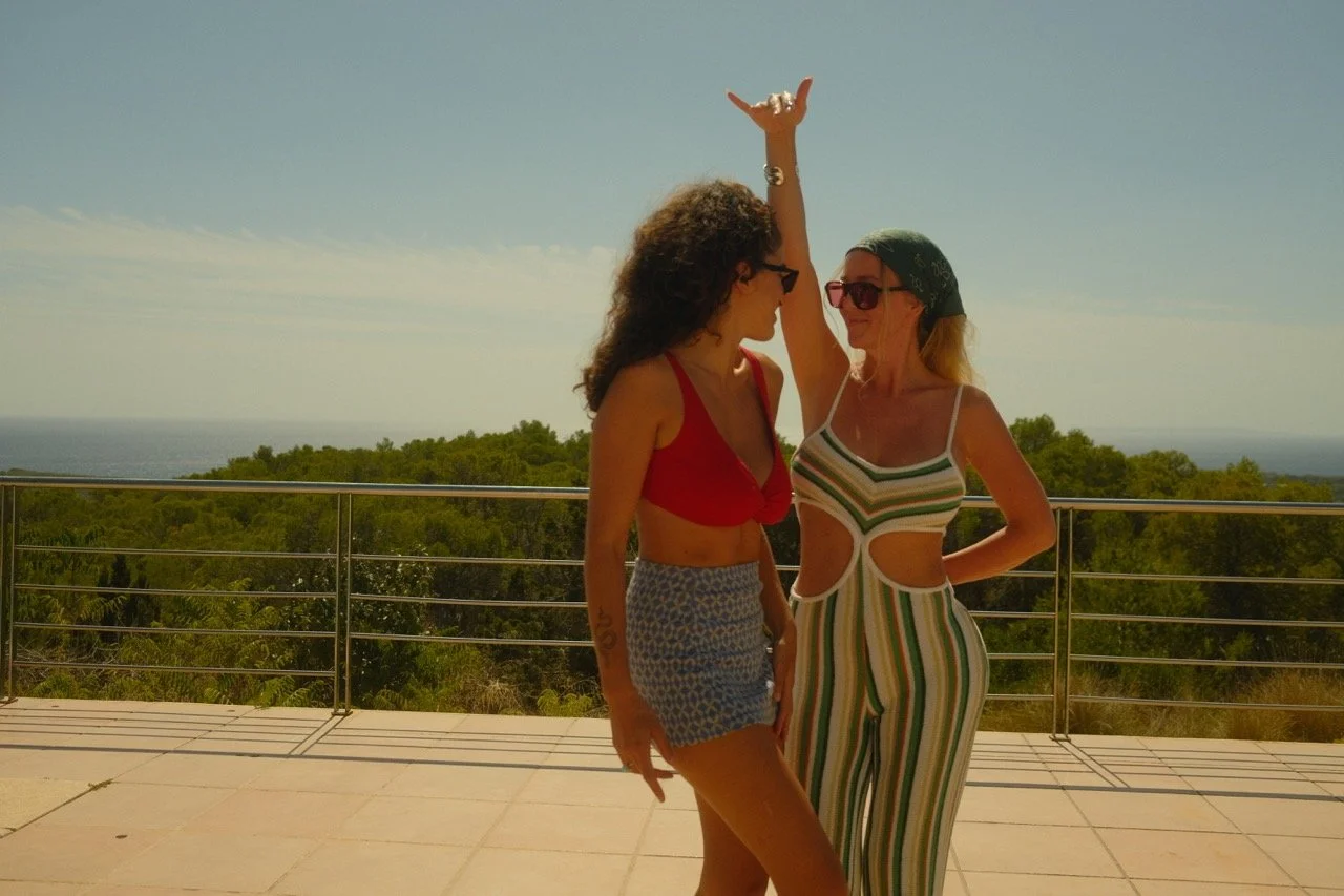 Zwei Frauen auf einer Terrasse im Sonnenlicht, umgeben von Natur, die lachen und tanzen.