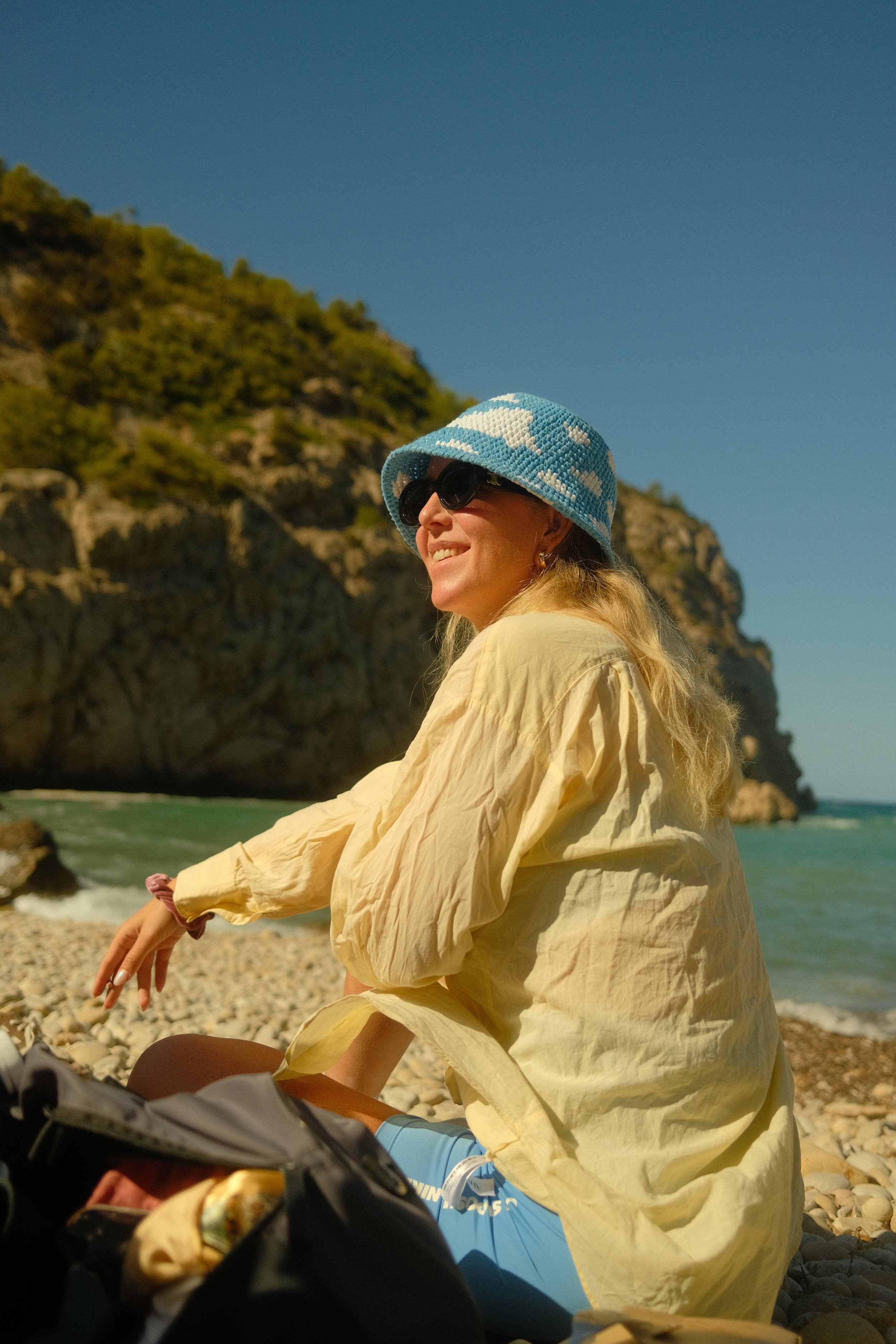 Frau mit Sonnenbrille, blauer Hut mit weißen Wolkenmustern und gelber Jacke sitzt am Strand mit Blick auf das Meer und Klippen im Hintergrund.