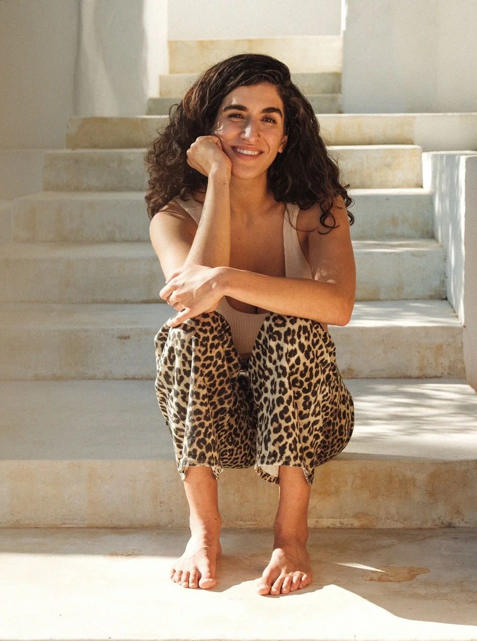 Lächelnde Frau mit dunklen, lockigen Haaren, sitzend auf weißen Treppen, trägt Leopardenmuster-Hose und beige Top.