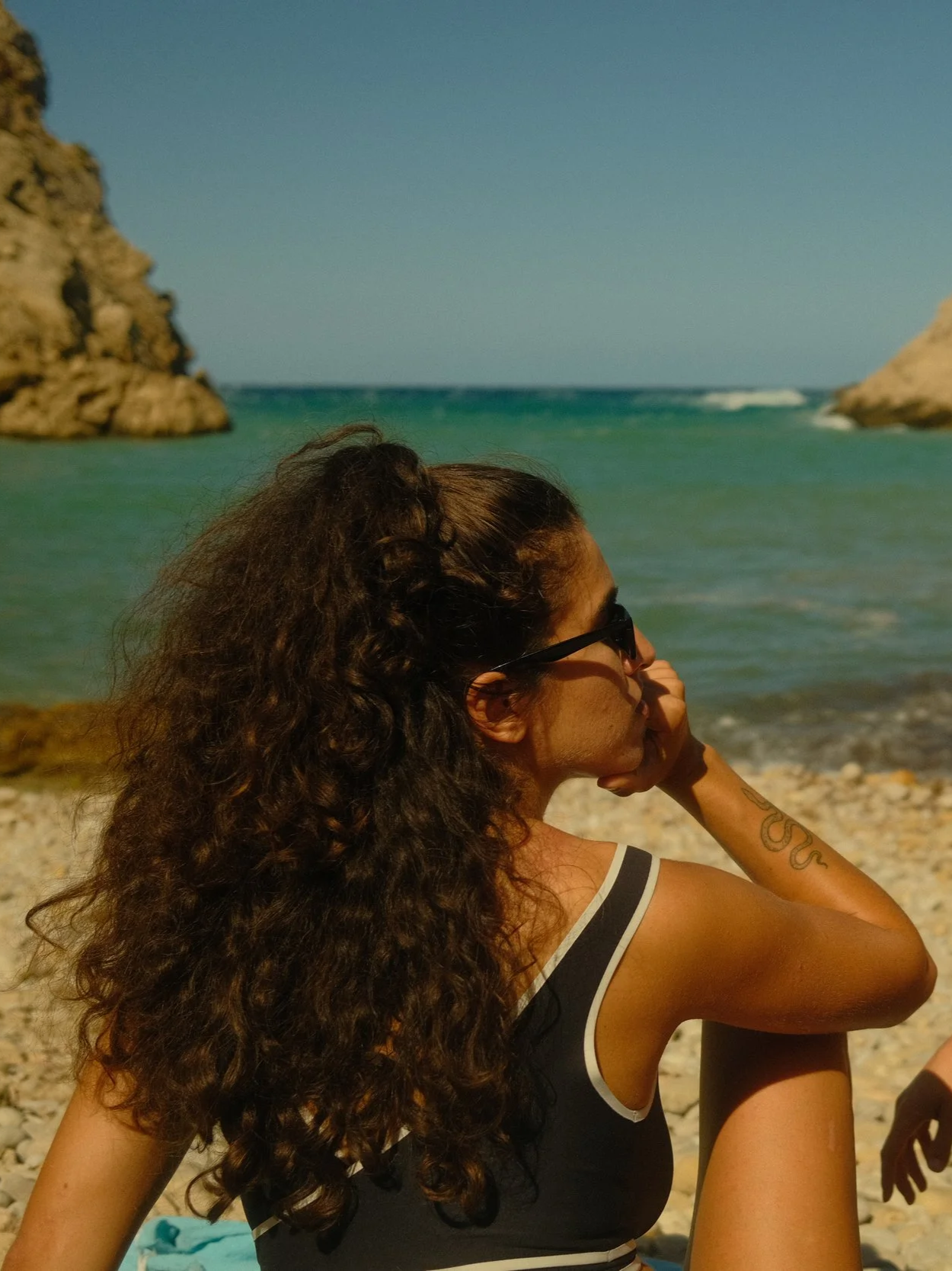 Junge Frau mit langen, lockigen Haaren und Sonnenbrille, sitzend am Strand, blickt aufs Meer, im Hintergrund Felsen und bewölkter Himmel.