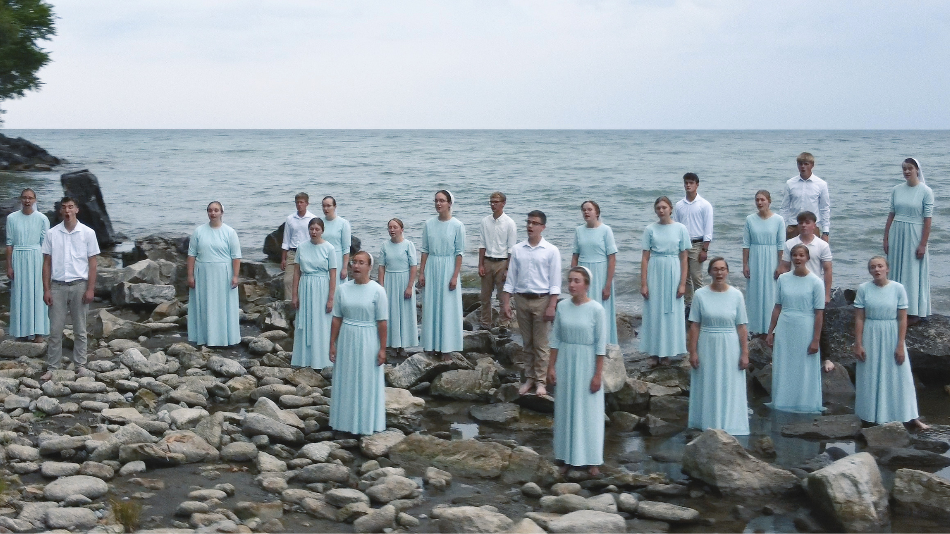 A choir of young people stand on rocks by the ocean, singing during daytime. The group includes both males and females, most dressed in light blue dresses or white shirts with khaki pants.