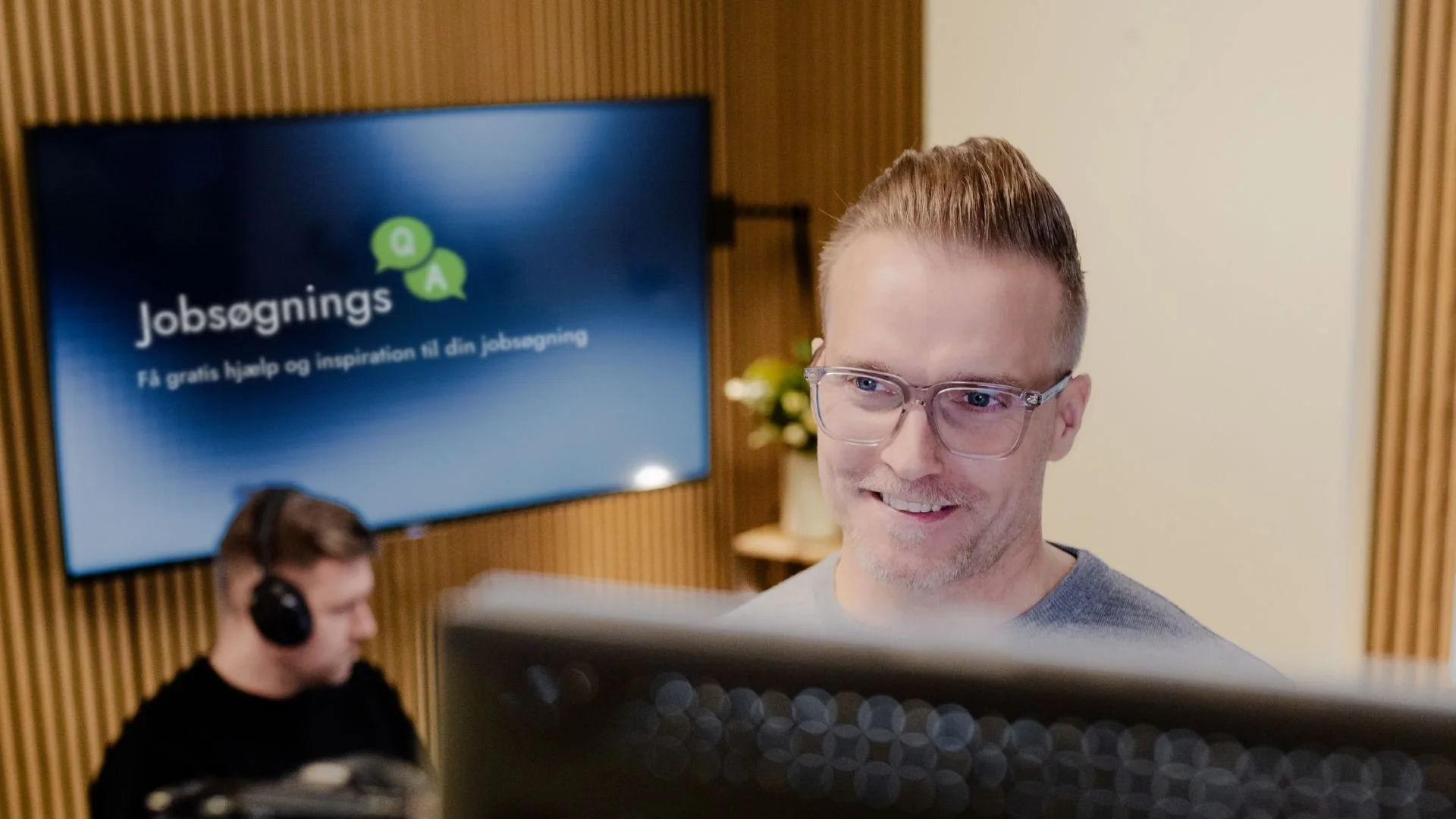 En mand med briller ser på en computerskærm, mens en mand med headset arbejder ved en computer i baggrunden. Skærmen viser teksten 'Jobansøgning'.