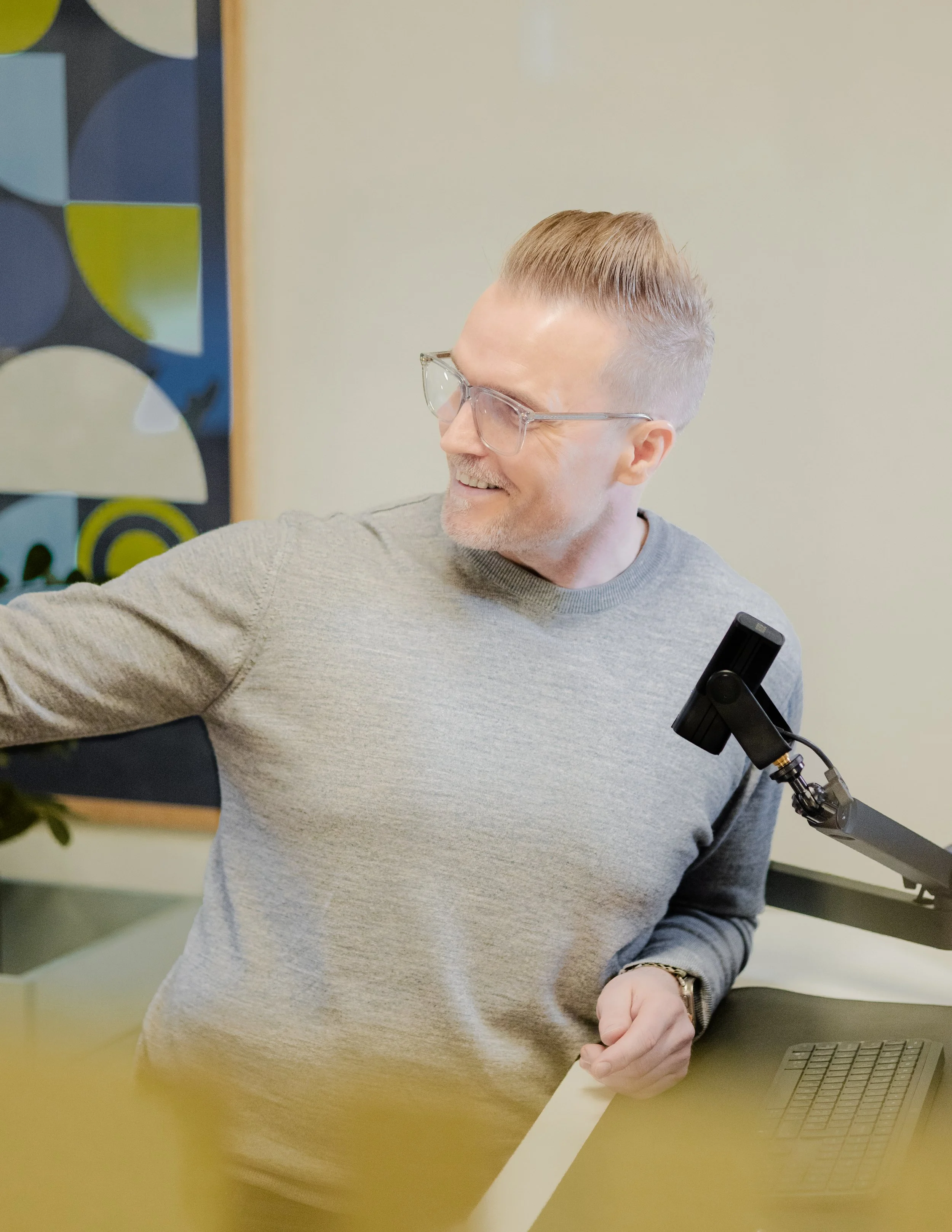 En mand med briller og gråt hår, smilende, iført en grå sweater, står foran en computer og peger på noget uden for billedet.