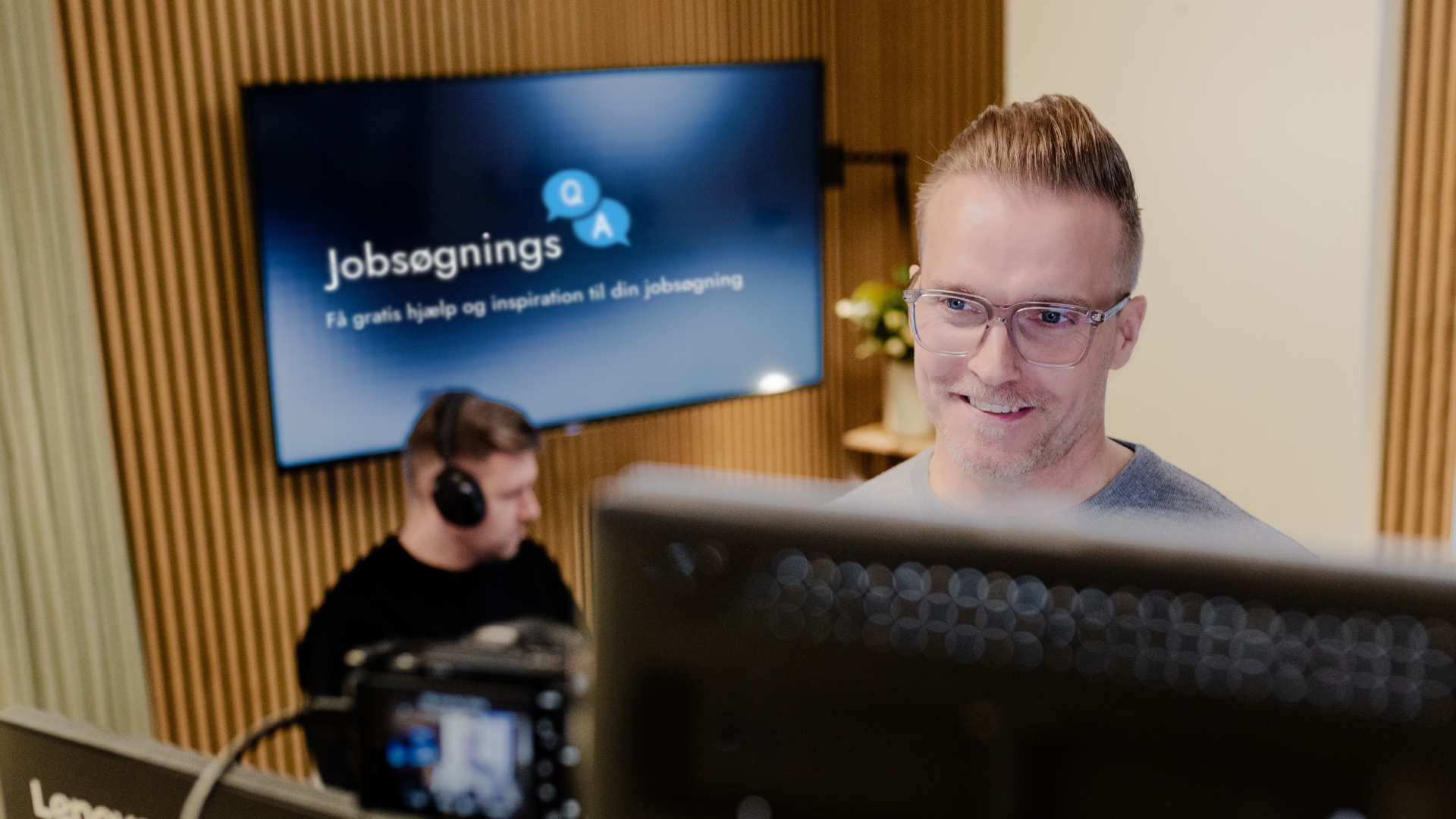 En mand med briller smiler foran en computerskærm under en digital optagelse med en baggrund med en stor skærm, der viser teksten 'Jobsøgnings' og 'Få gratis hjælp og inspiration til din jobsøgning', mens en anden person sidder med hovedtelefoner.