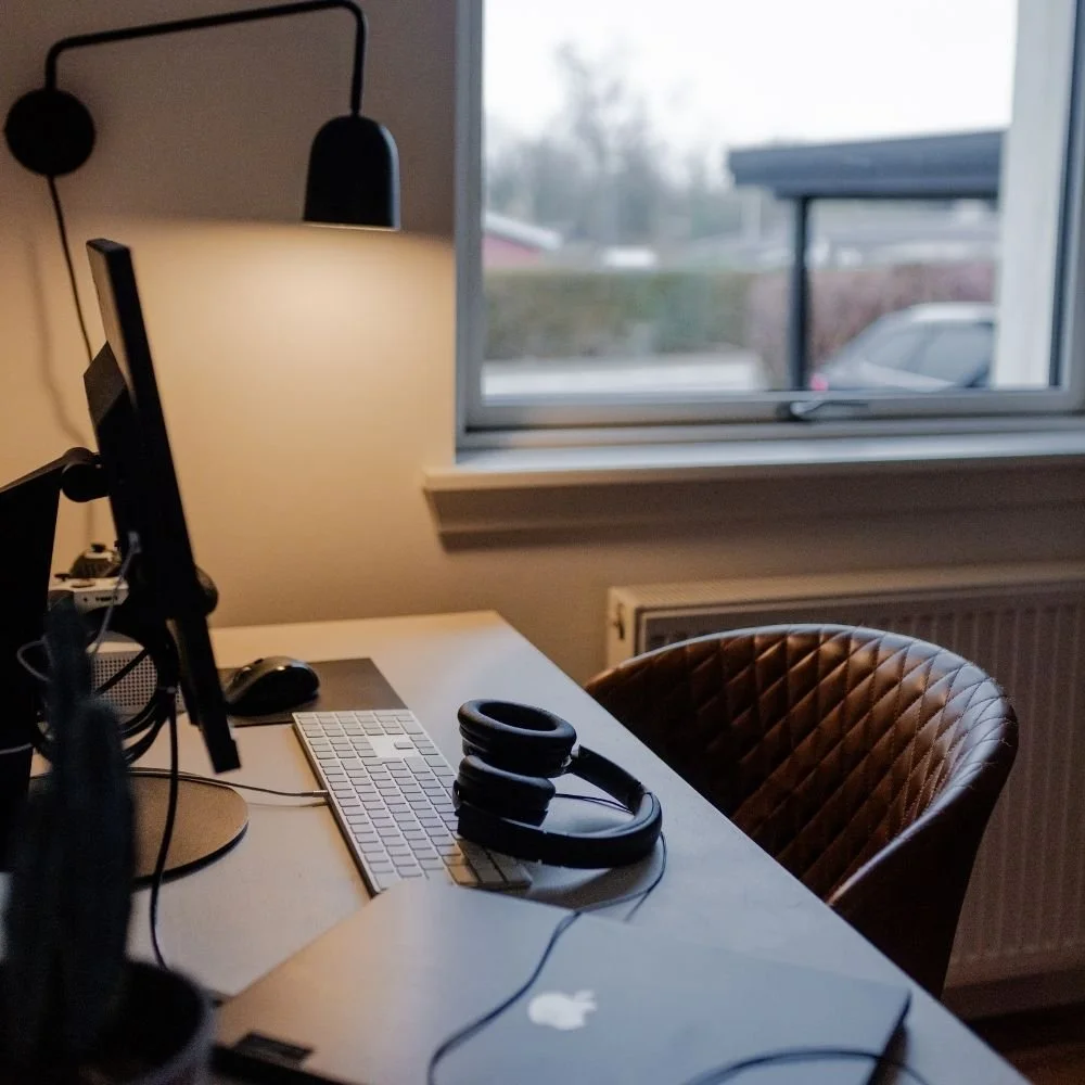 Arbejdslampe, computer, tastatur, mus og hovedtelefoner på et skrivebord ved et vindue i et hjemmekontor.