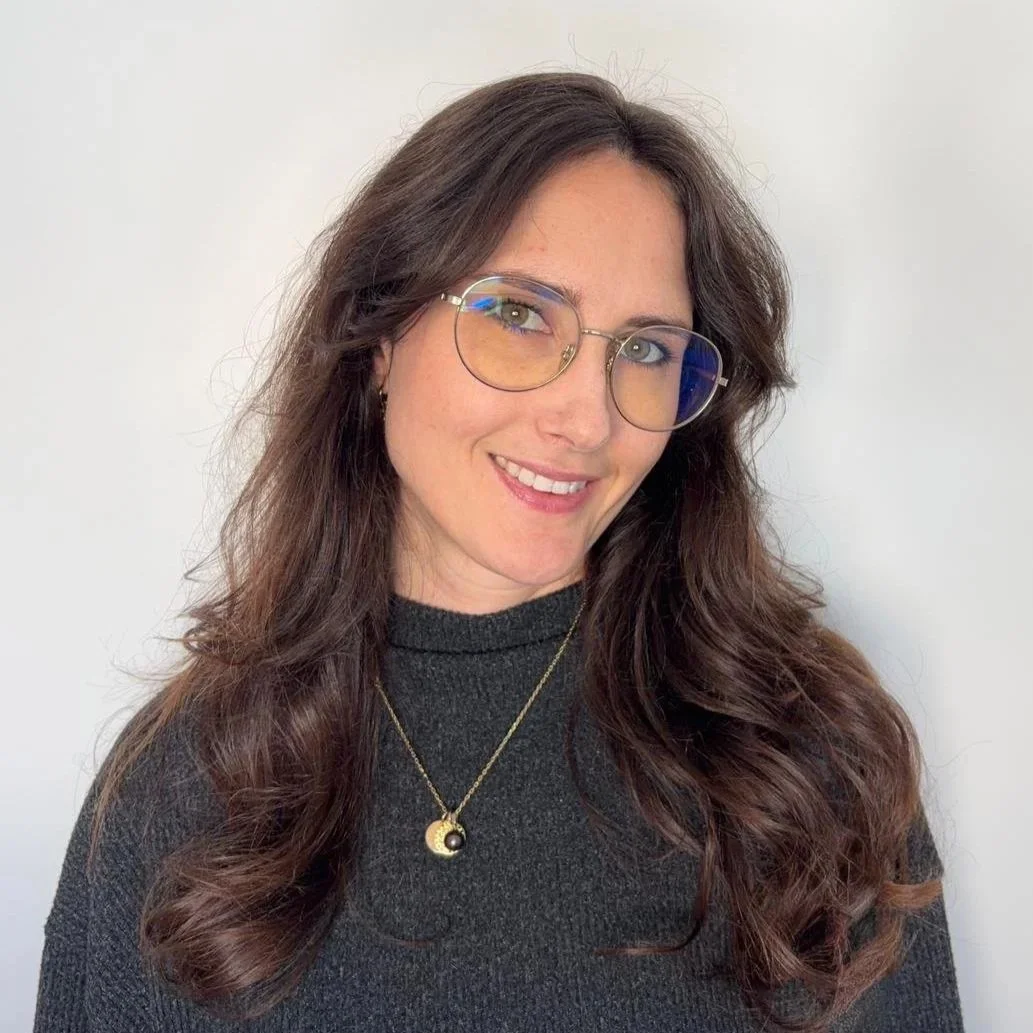 Retrato de una mujer con cabello castaño oscuro, lentes con montura metálica, luciendo una sonrisa y usando una camisa gris oscuro con un collar dorado. Fondo blanco.