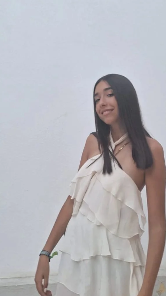 Joven con cabello largo, vestido blanco con volantes, sonriendo, en un fondo blanco.