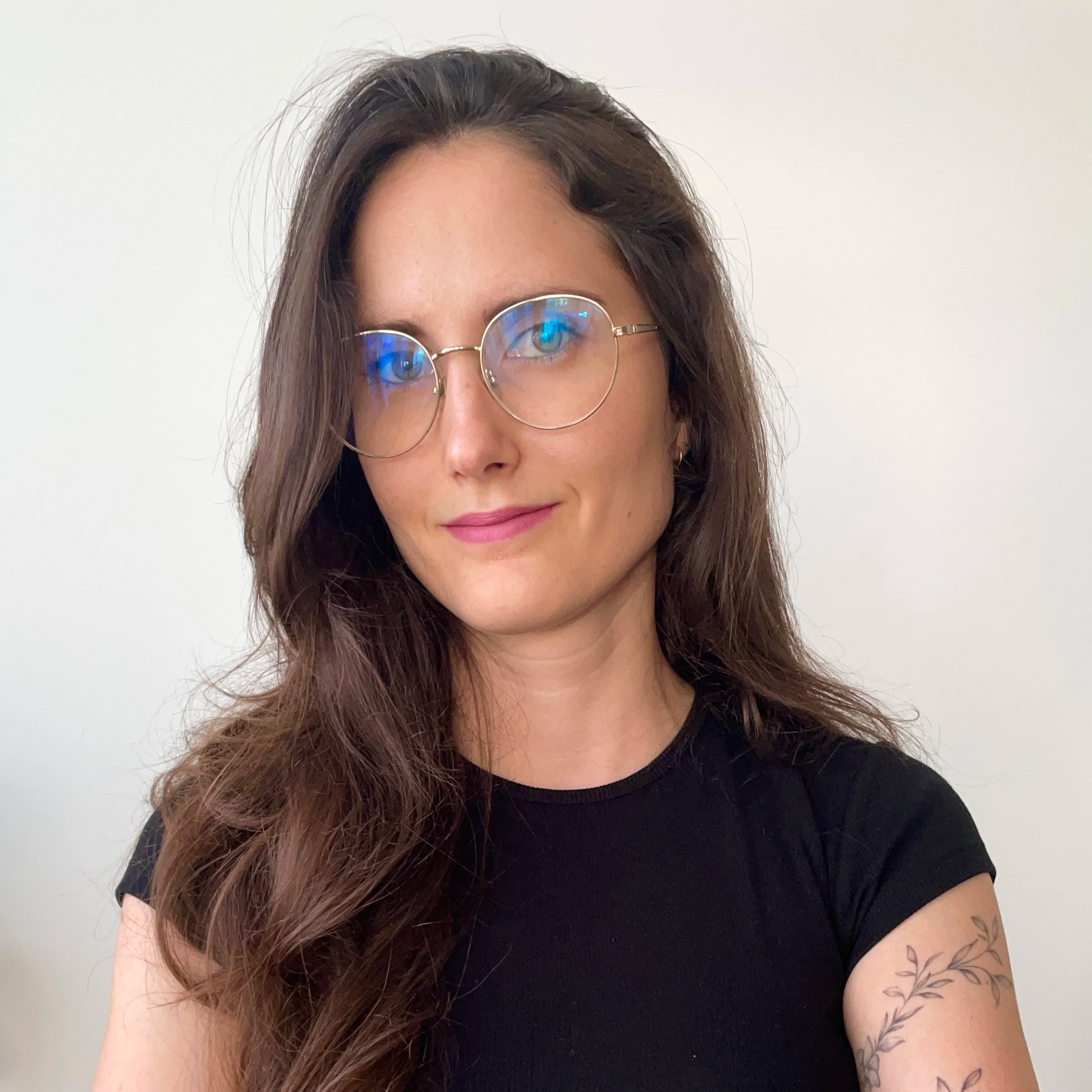 Mujer con gafas redondas, cabello largo y rizado, camiseta negra, tatuaje en brazo, fondo blanco