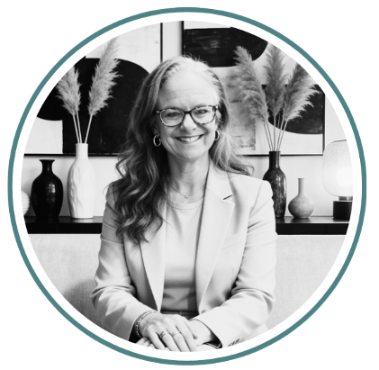 A woman with glasses and long hair smiling, sitting at a table with decorative vases and plants in the background.