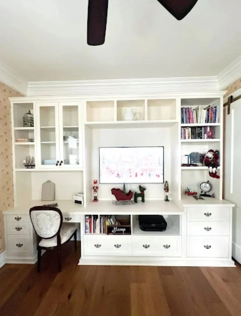 White built-in wall unit with shelves and drawers, decorated with Christmas-themed items, a computer monitor, books, and a small chair in front of the desk, in a room with hardwood floors.