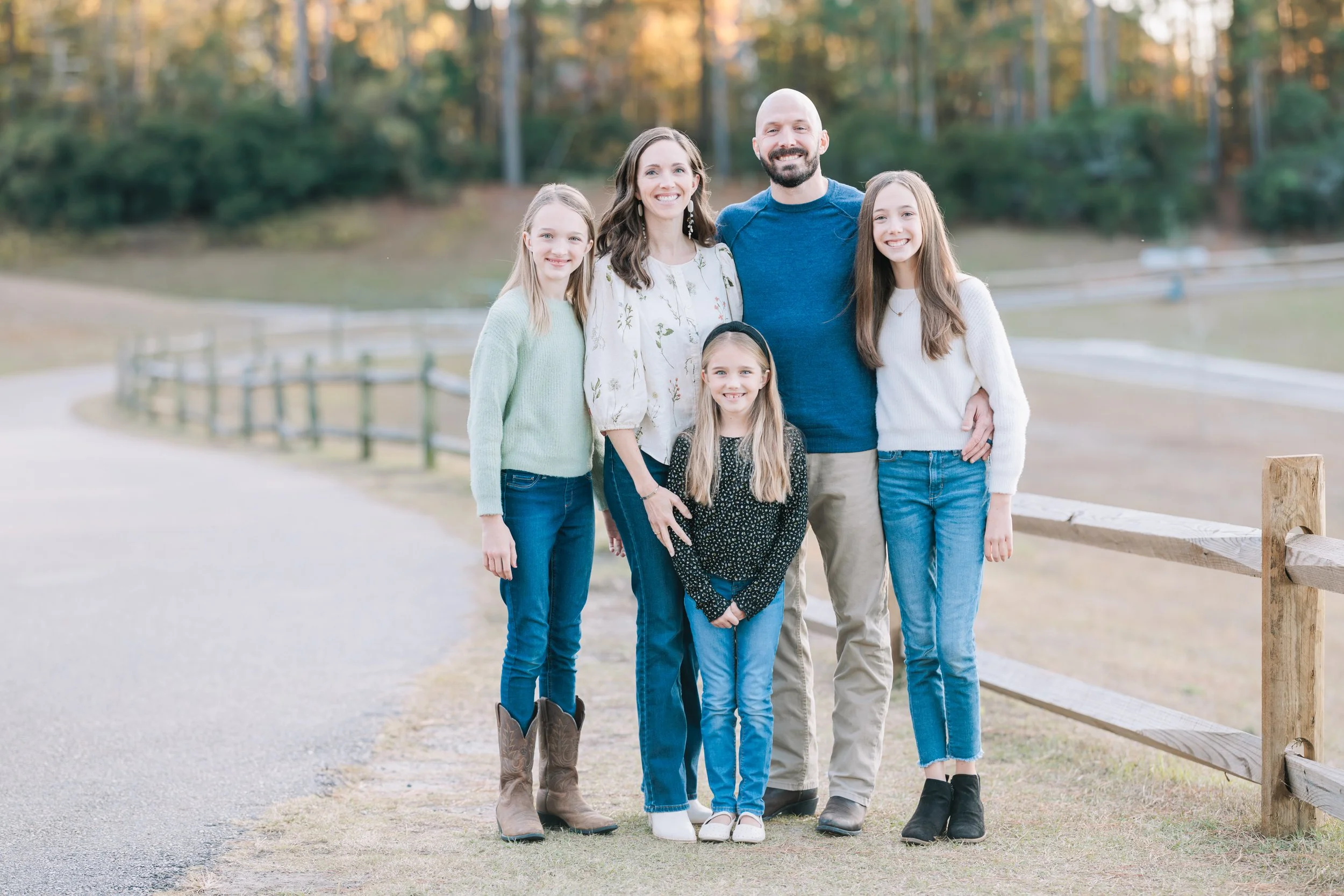 A family of six, including three children and two adults, standing outside on a walking path with a wooden fence and trees with autumn foliage in the background, smiling at the camera.