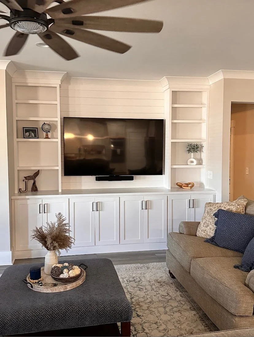 Living room with wall-mounted flat-screen TV, built-in white cabinets with open shelves, beige sofa with pillows, dark upholstered ottoman with decorative tray, vase with dried flowers, and ceiling fan.