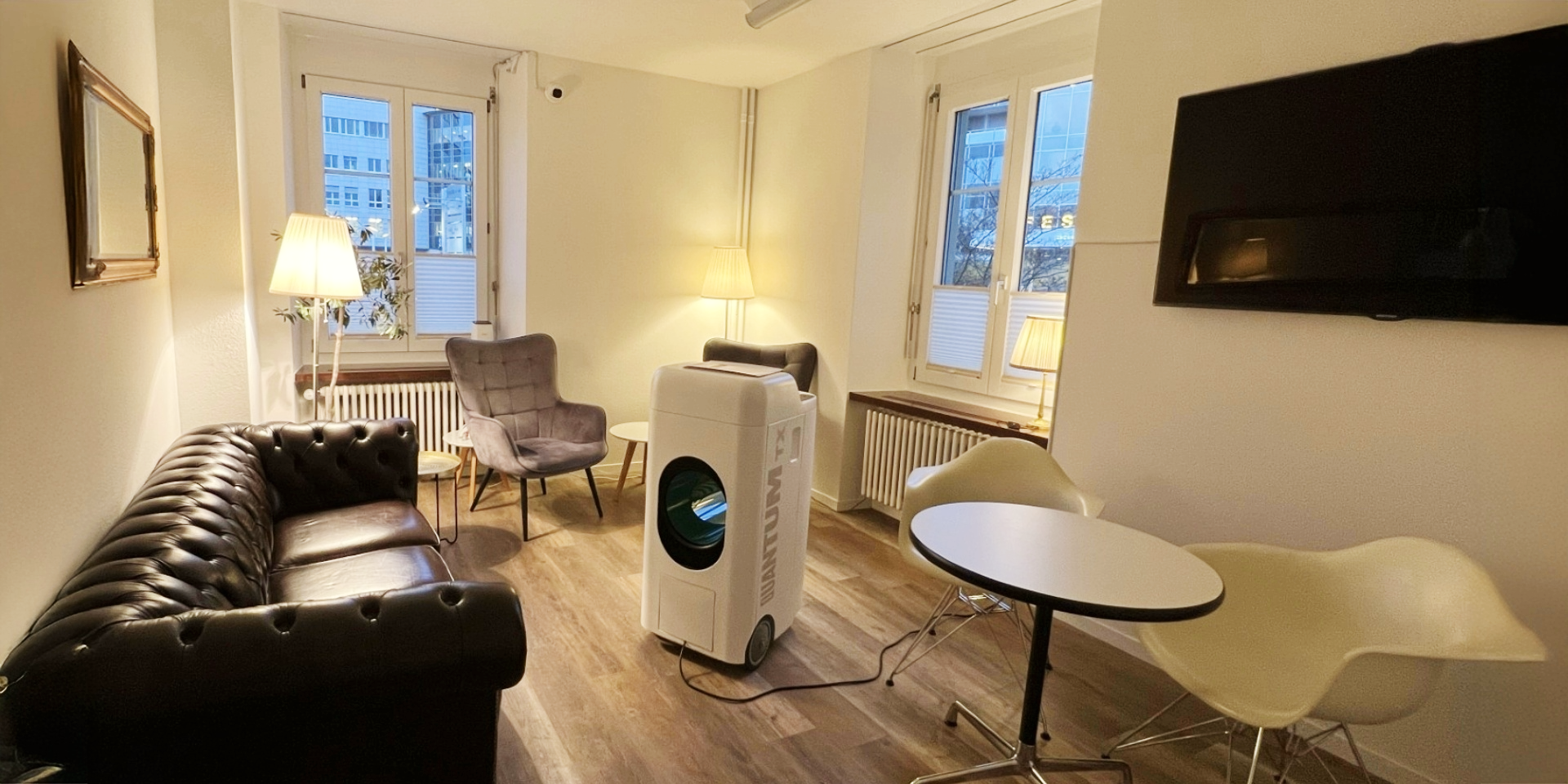 Ein modern eingerichteter Wohnzimmerraum mit braunem Ledersofa, mehreren Sessel, Stehlampen und einem Fernseher. Es gibt Fenster mit Blick auf ein Stadtbild, und in der Mitte steht ein Luftentfeuchter.
