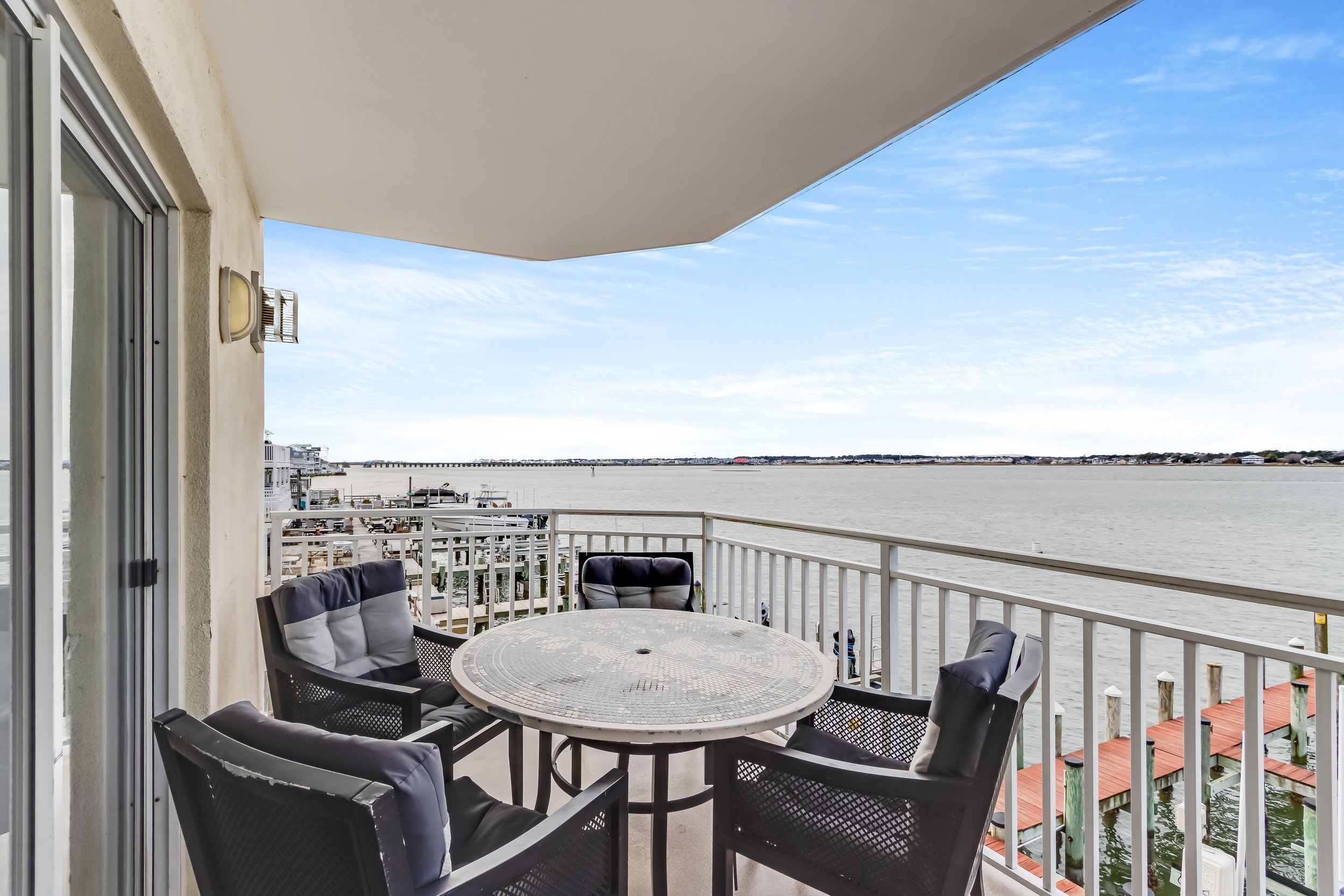 Balcony overlooking a body of water with a round table and four chairs, some with cushions, railing, and neighboring buildings in the distance under a partly cloudy sky.