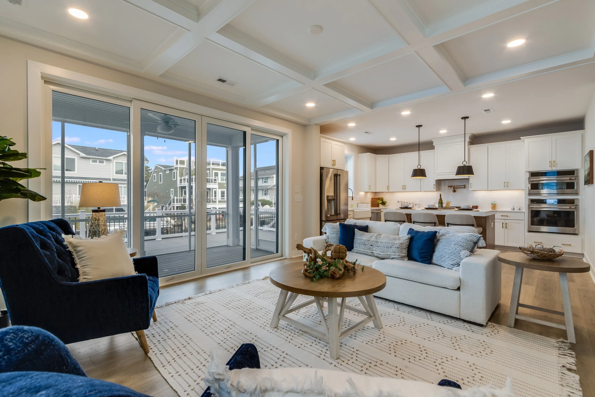 A modern open-concept living room and kitchen with large glass sliding doors leading to a balcony, featuring white cabinets, stainless steel appliances, a kitchen island with bar stools, and a cozy seating area with blue and white pillows, a white ru