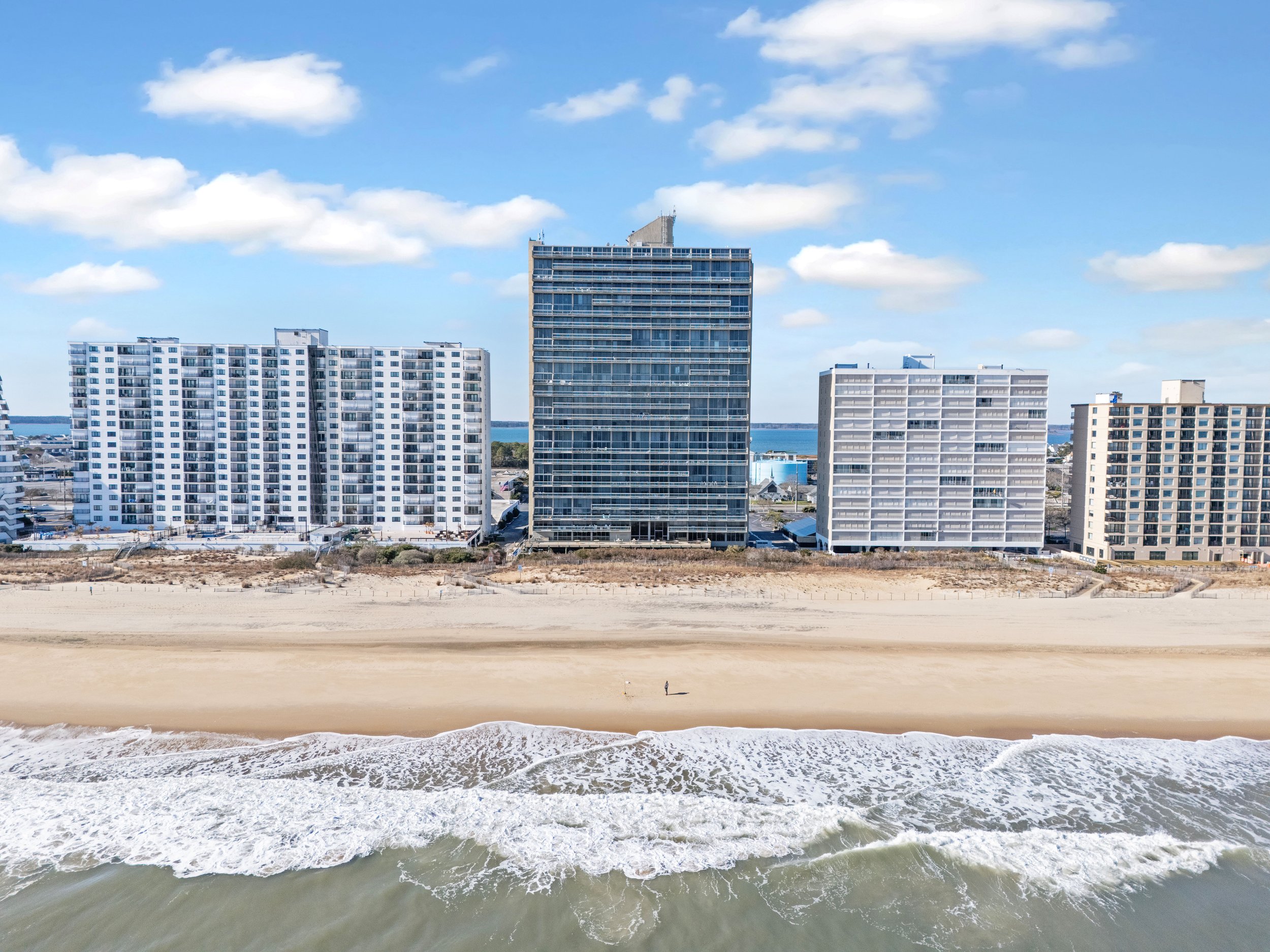 Beach with waves in the foreground and a row of high-rise buildings along the shoreline, with a blue sky and scattered clouds.