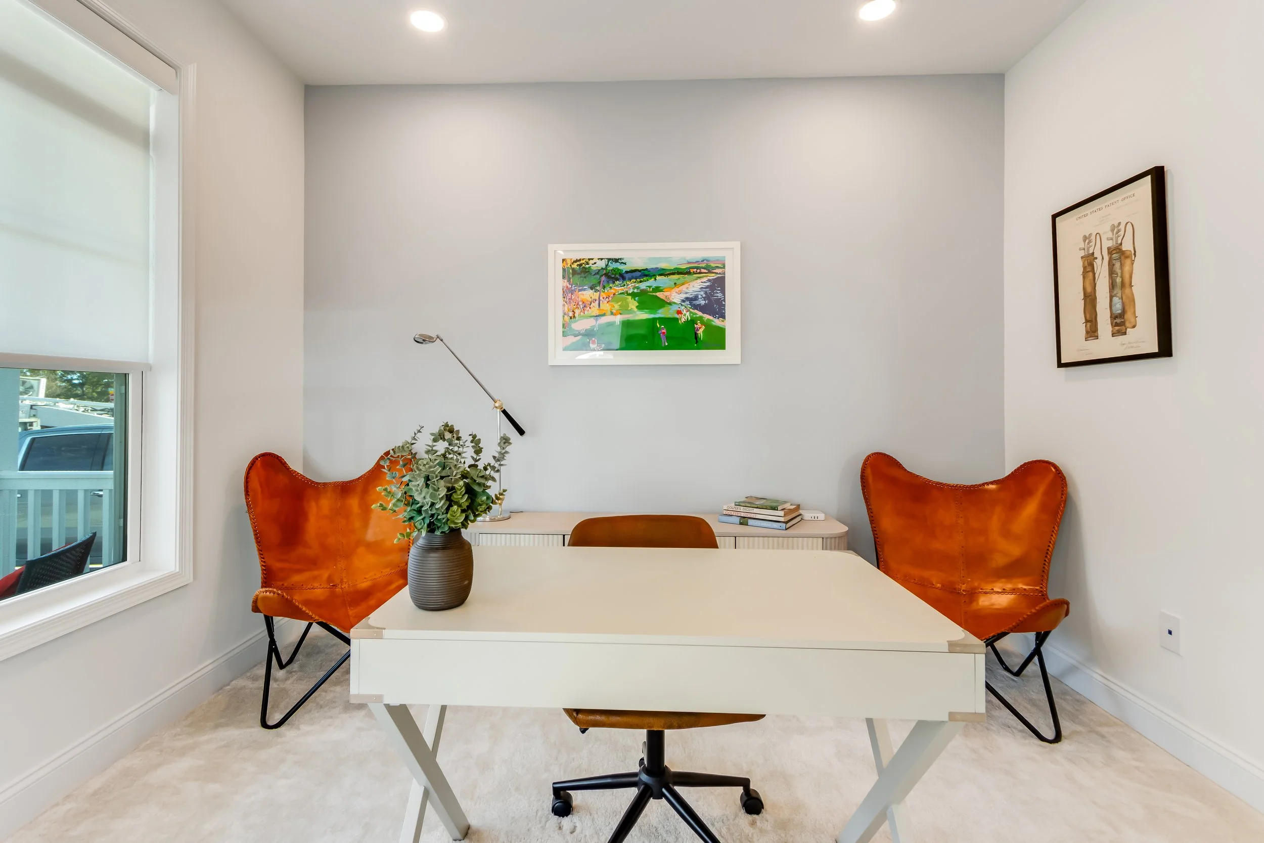 A modern home office with a white desk, an office chair, two orange leather lounge chairs, a window with a blind, a potted plant, framed artwork on the walls, and books and a lamp on a sideboard.