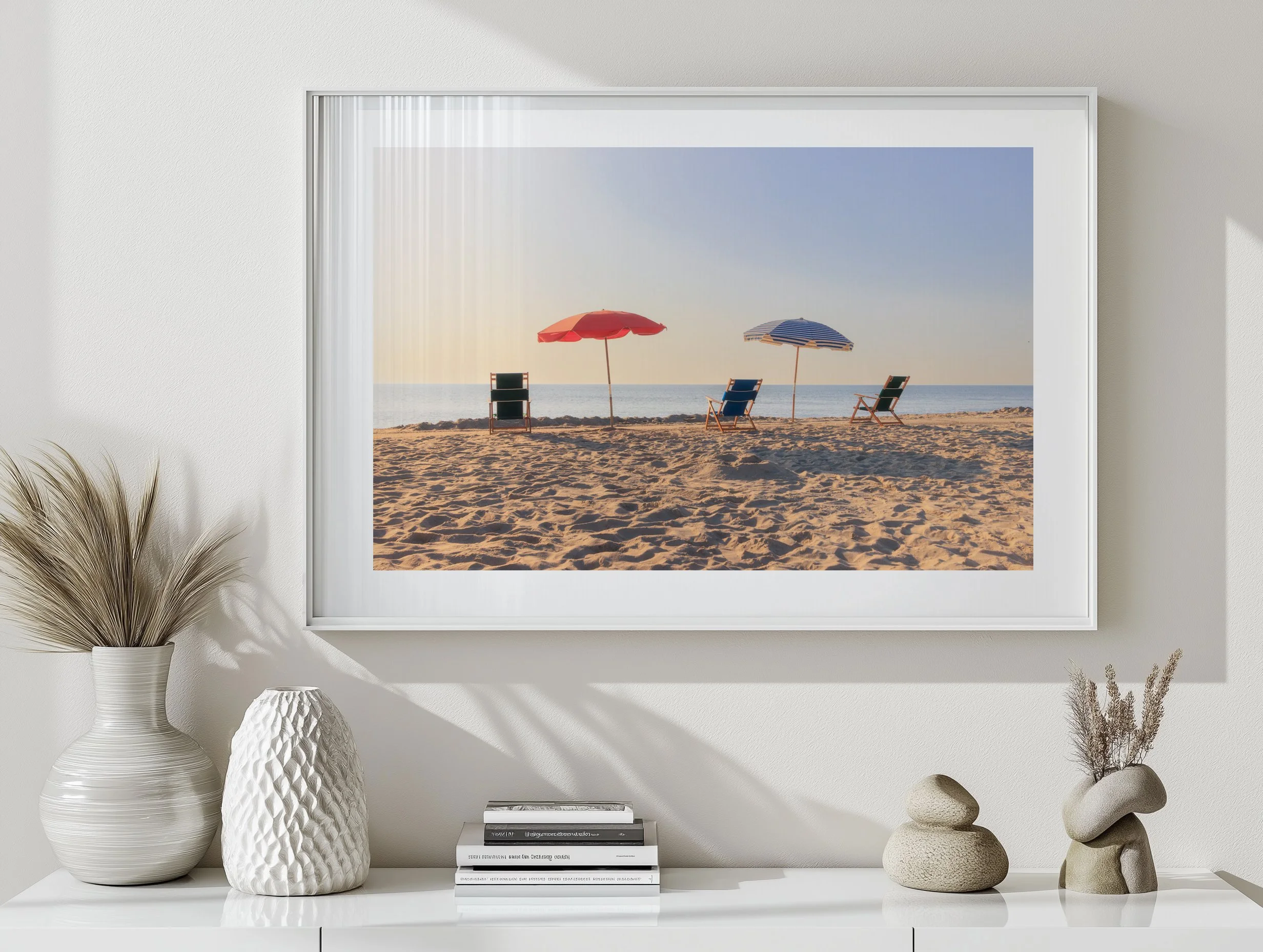 Framed photograph of a sandy beach with three lounge chairs, two umbrellas, and the ocean in the background, hung on a white wall.