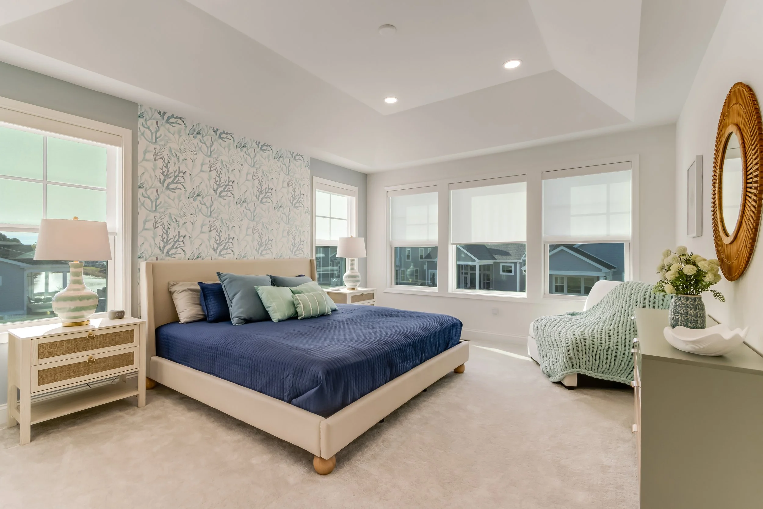 A bedroom with a beige bed, blue blanket, multiple pillows, two nightstands with lamps, three large windows, a white armchair with a knitted blanket, and a dresser with a vase of flowers.