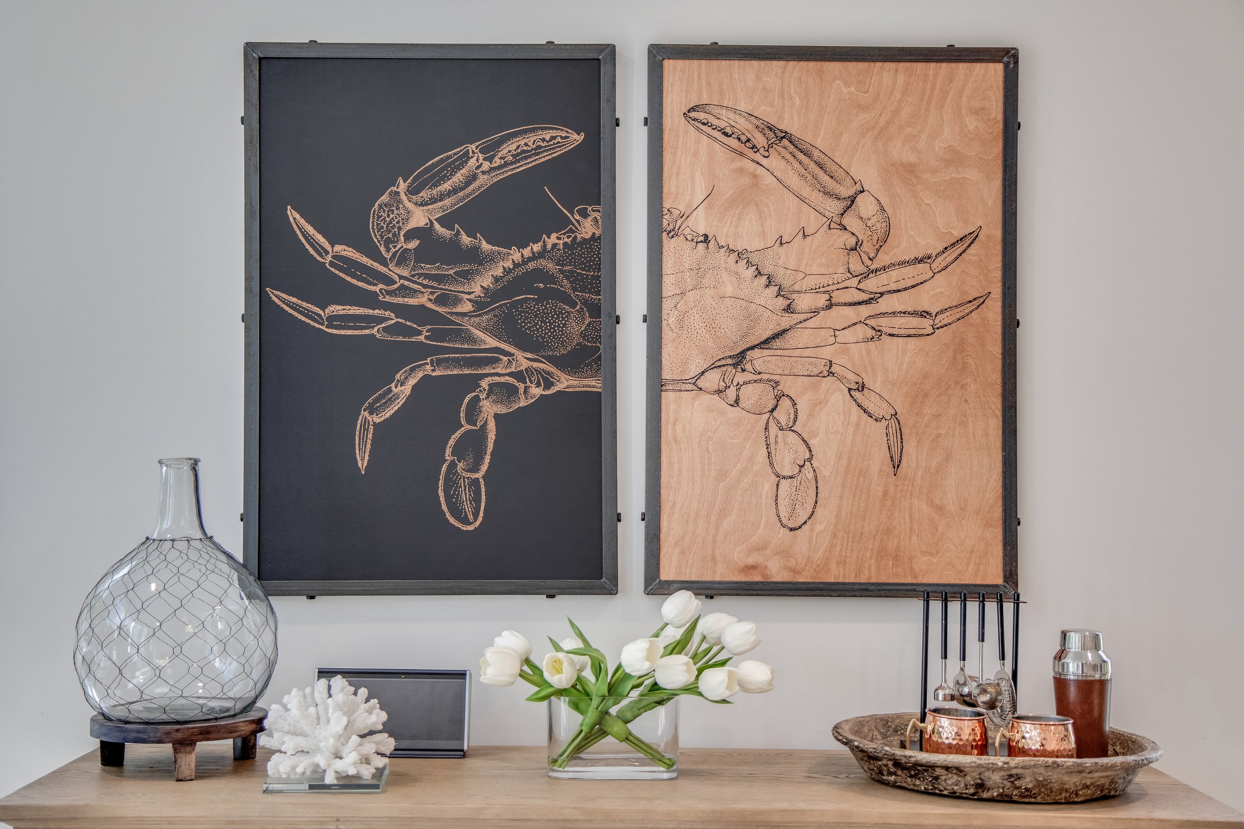 Decorative wall art featuring black and wood panels with lobster illustrations, placed above a wooden table. The table displays a large glass vase with a wire mesh design, a piece of white coral, a glass container of flowers, and a tray holding a var