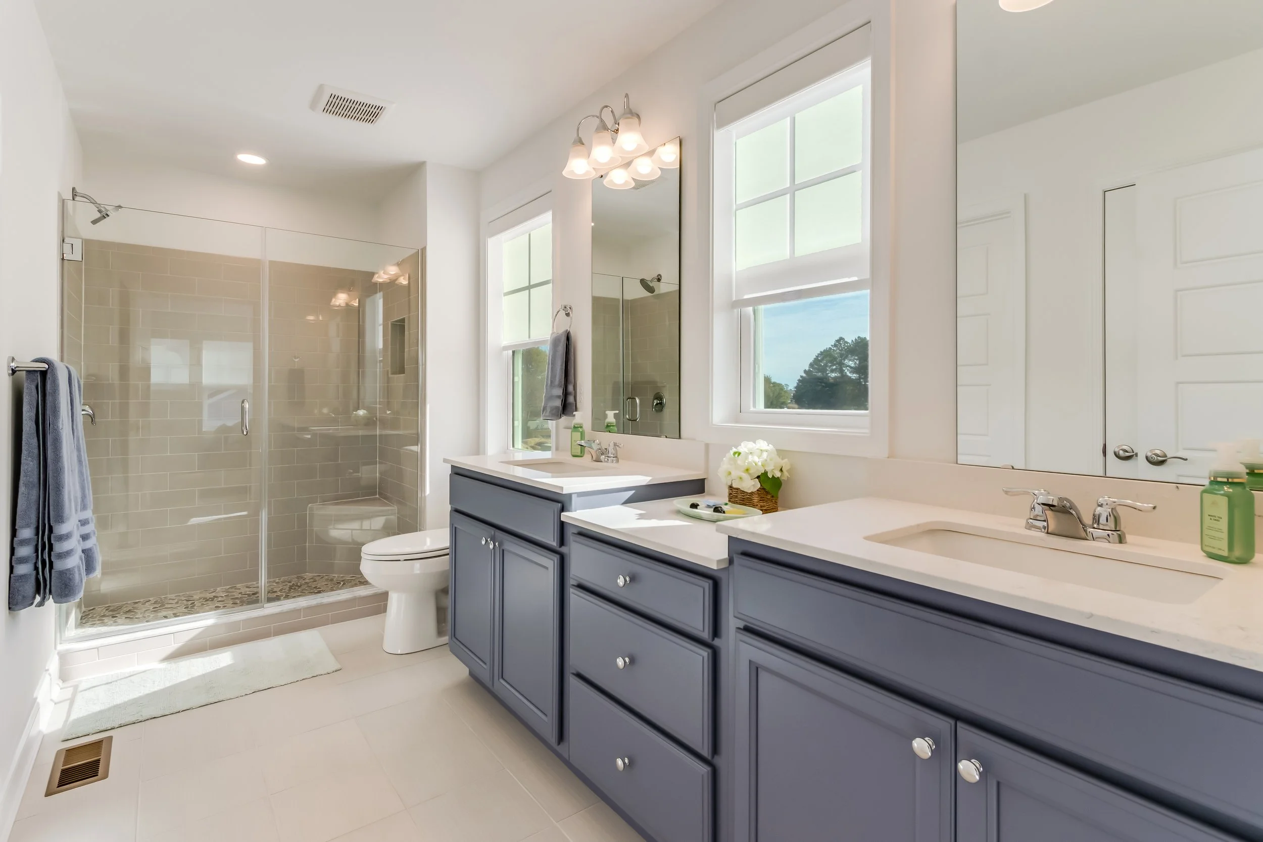 Bright bathroom with double vanity sinks, navy blue cabinets, large mirror, window with a view of trees, glass-enclosed shower with beige tiles, towel on the rack, and small decorative flowers on the countertop.