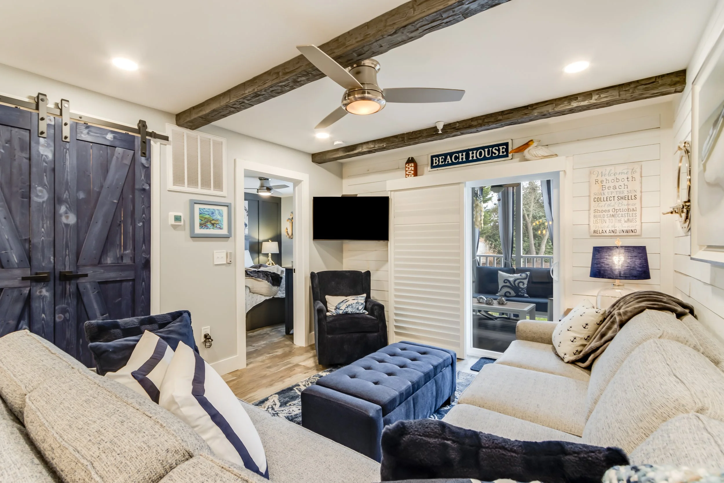 Cozy living room with white shiplap walls, wooden ceiling beams, a sliding glass door leading to a porch, a beige sofa with nautical pillows, a black armchair, a blue ottoman, a wall-mounted flat-screen TV, and beach-themed decor including a sign that says 'Beach House'.