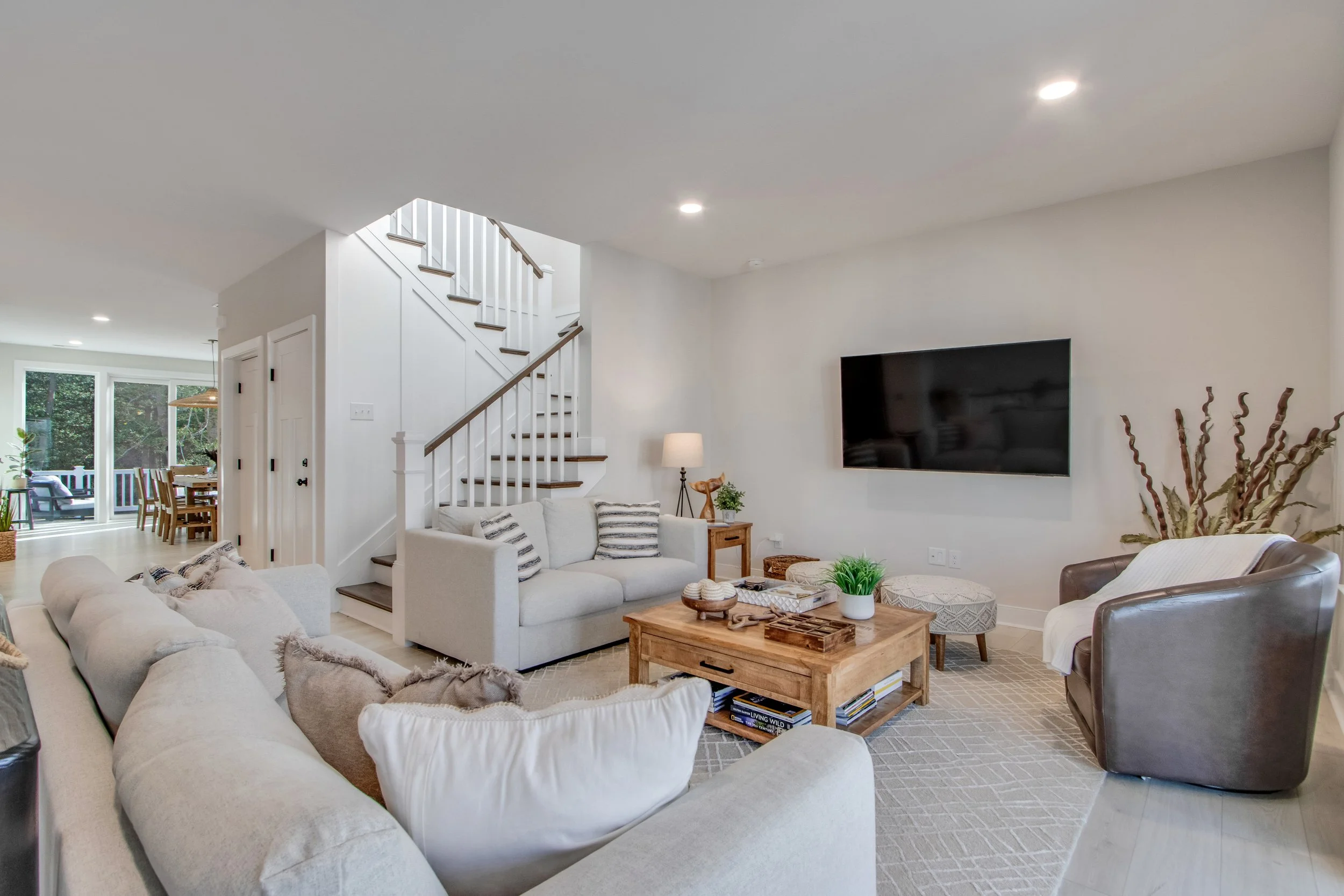 Bright living room with white walls, beige sofas, a wooden coffee table, a mounted flat-screen TV, a leather armchair with a white throw, and a staircase with white risers and wooden treads leading to the upper floor.