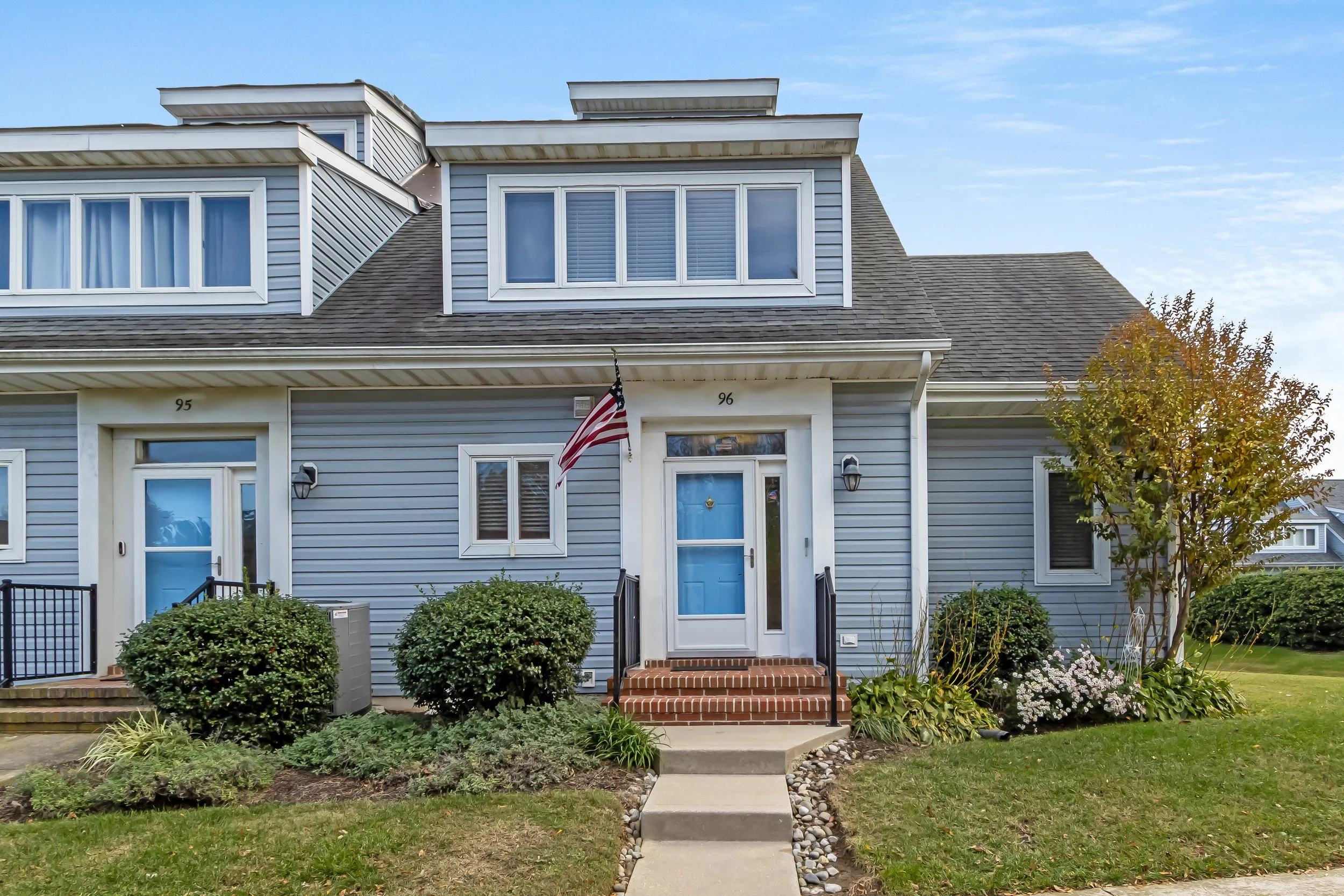 Front view of a blue townhouse with white trim, featuring a small set of brick stairs, an American flag, and landscaped garden with bushes and trees.