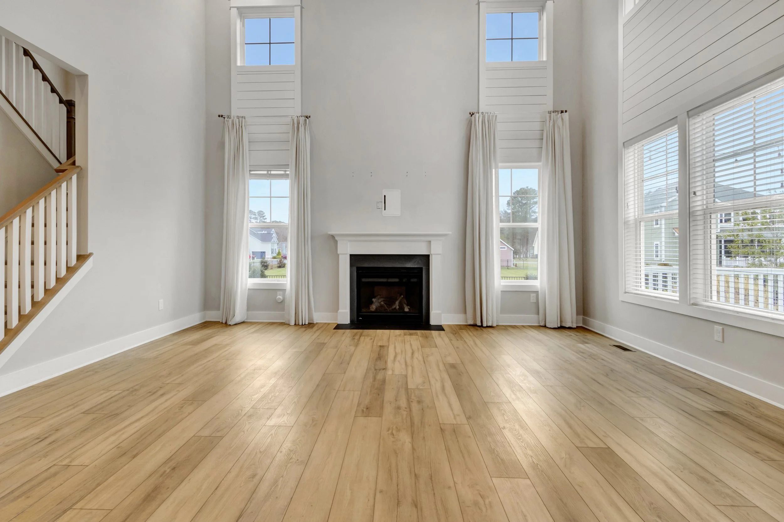 Empty living room with large windows, white curtains, a white fireplace, and hardwood floors.