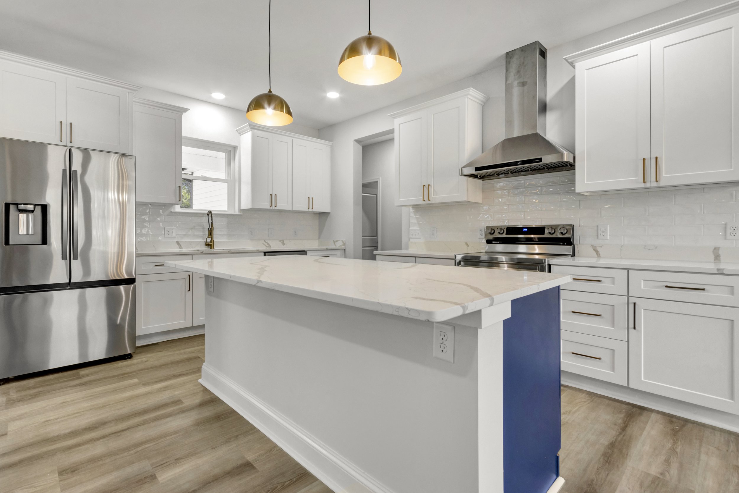 Modern white kitchen with stainless steel appliances, a central island with a marble countertop, pendant lighting, and wood flooring.