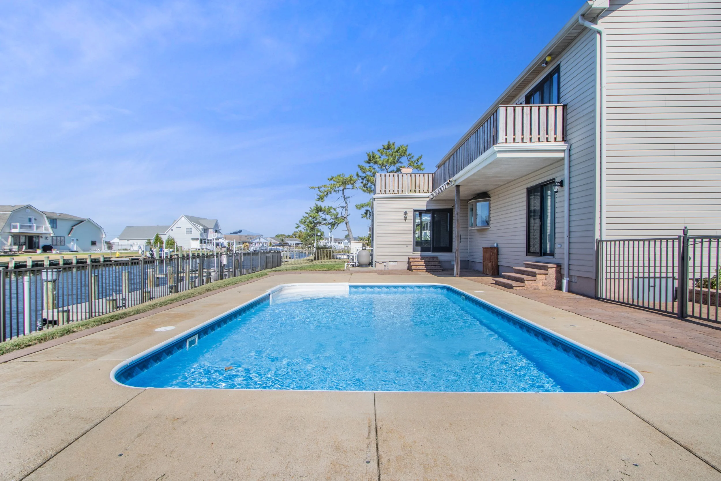 Residential backyard with a swimming pool, patio area, and house with balcony, overlooking water and neighboring houses under a clear blue sky.
