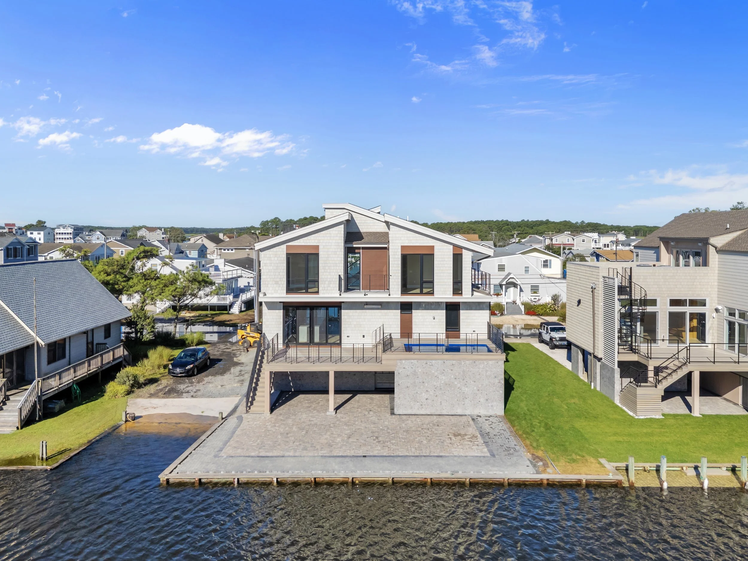Modern waterfront house on a grassy lot with a deck, overlooking a body of water, with neighboring houses in the background under a blue sky.