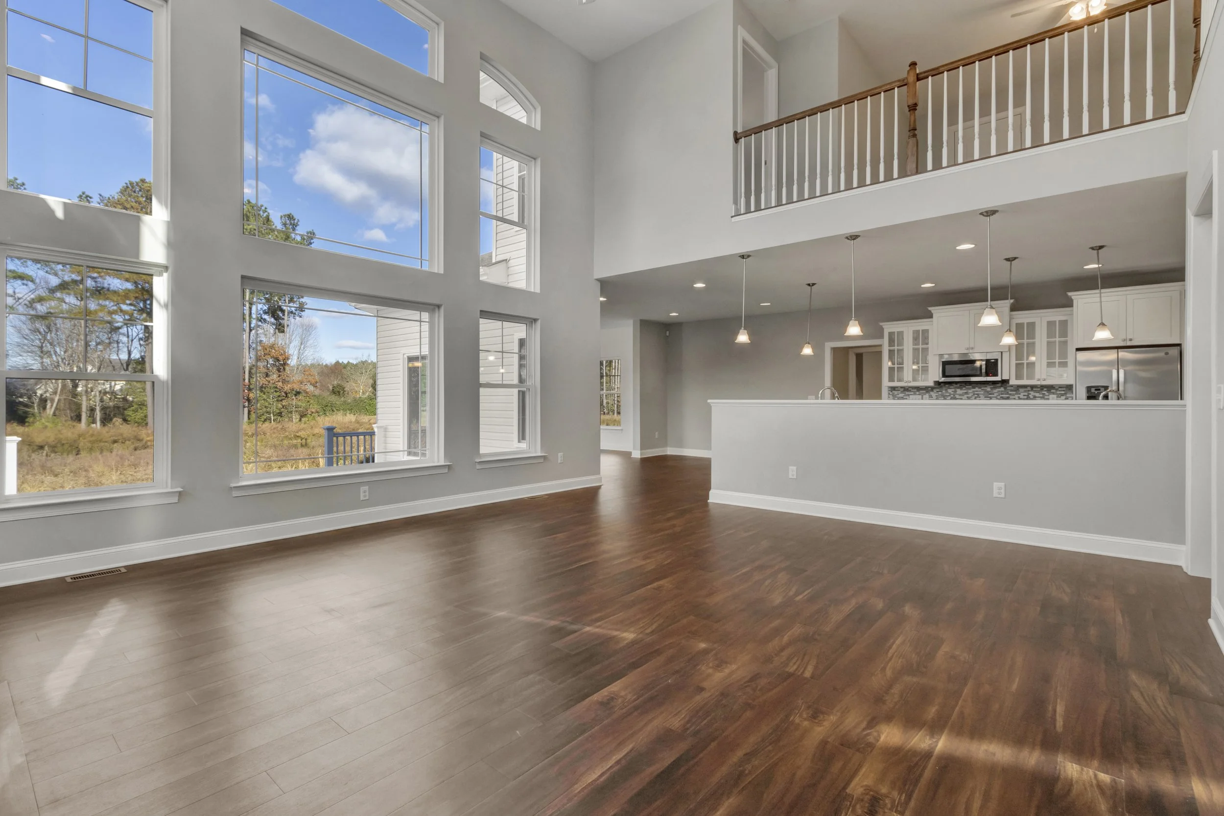 Empty living room with large windows, hardwood floors, and open kitchen with white cabinets and island