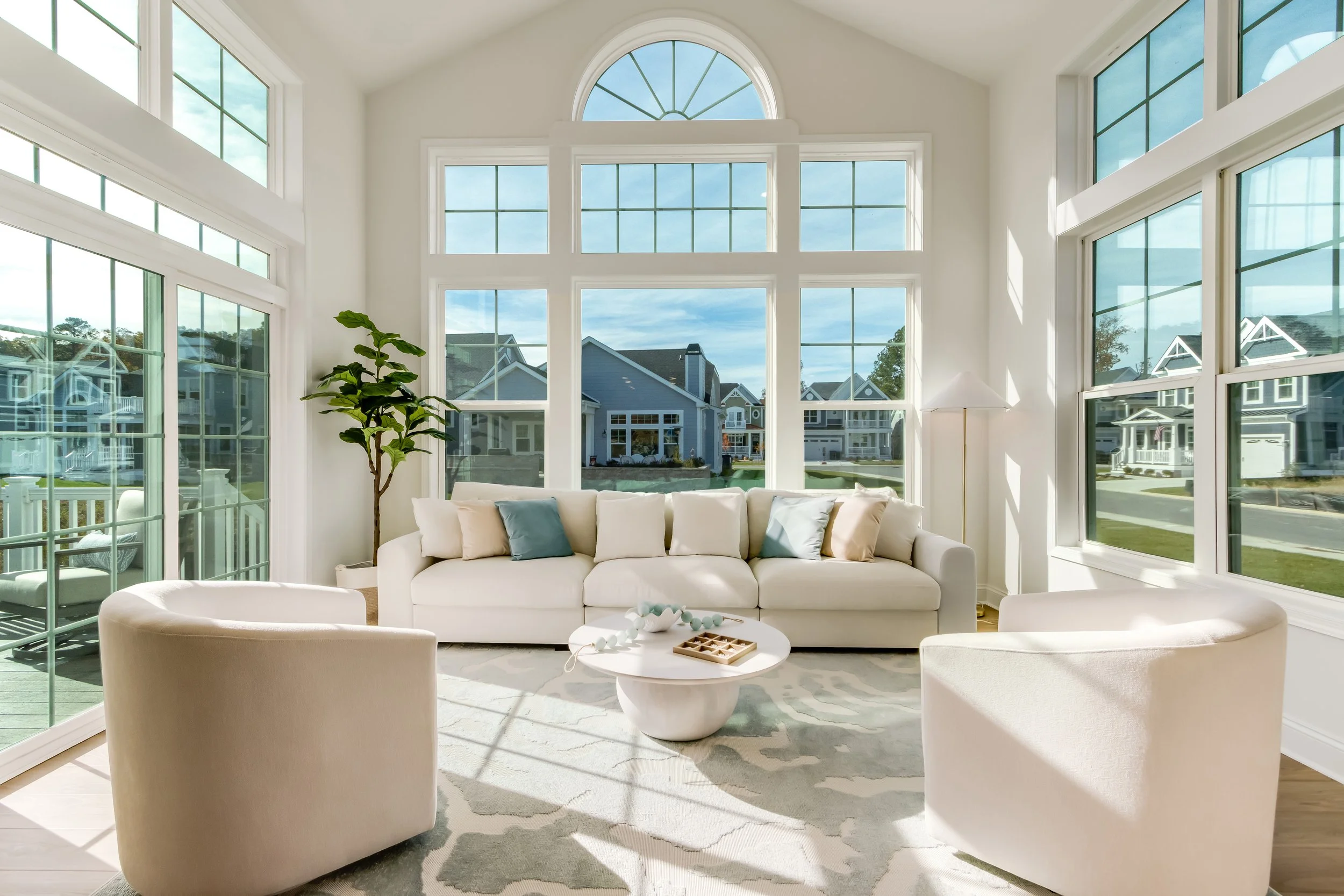 Bright living room with large windows, white sofa, two cream armchairs, round coffee table, green plant, and sunlight streaming in.