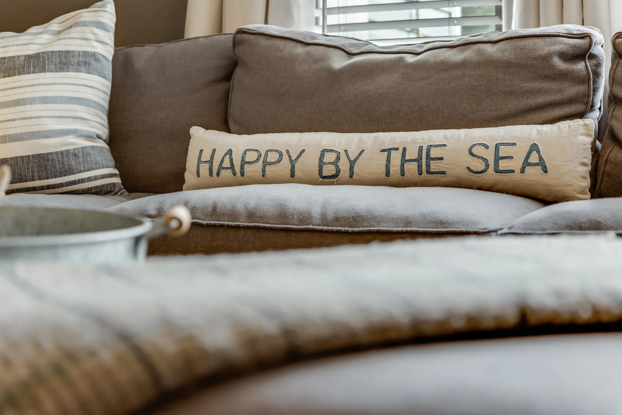 Decorative beige pillow with dark blue embroidery that reads 'HAPPY BY THE SEA' on a beige couch, with striped pillows and a window in the background.
