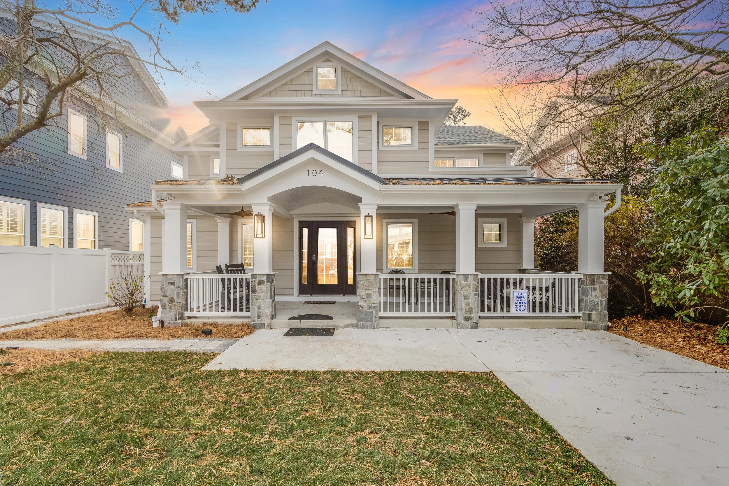 Front view of a new two-story house with a covered porch, stone pillars, black front door, and surrounding trees with some leaves fallen, during sunset.