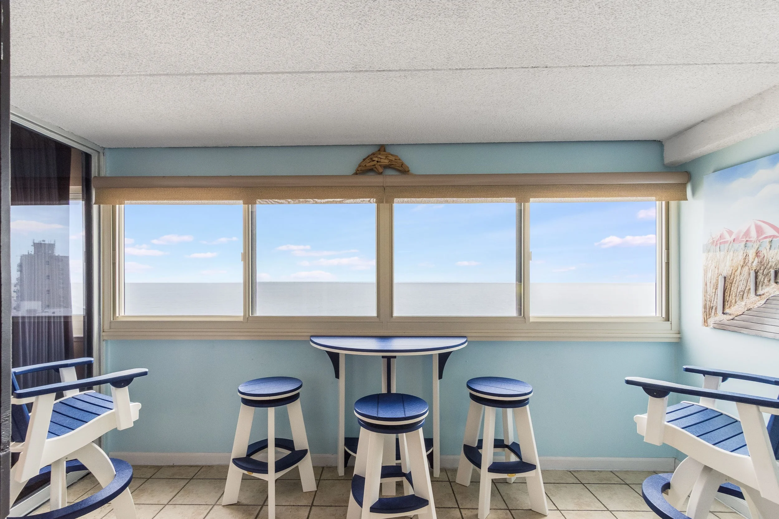 Indoor space with a view of the beach and ocean through large windows, featuring blue and white furniture including stools and chairs.