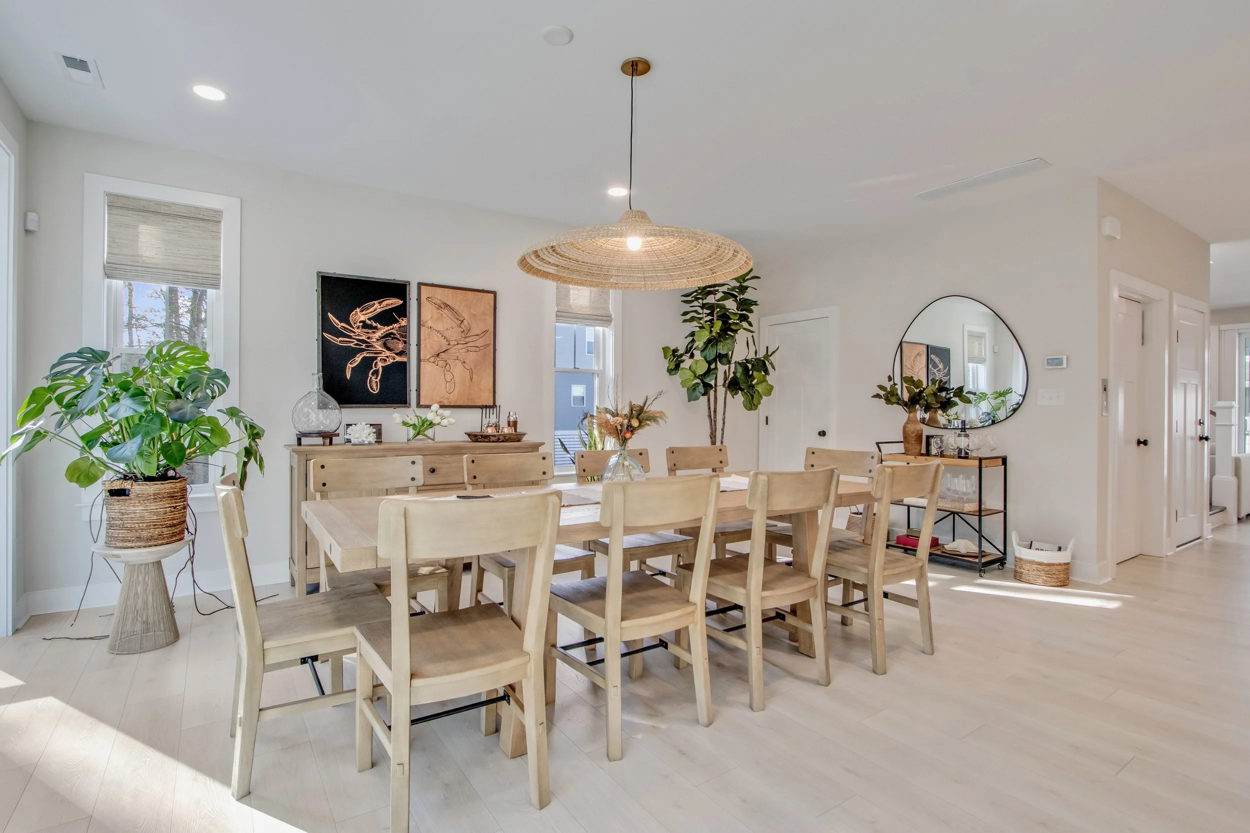 Bright dining room with a large wooden table, ten matching chairs, potted plants, a sideboard with artwork, and a wall mirror, illuminated by a woven ceiling light, with natural light coming through windows.