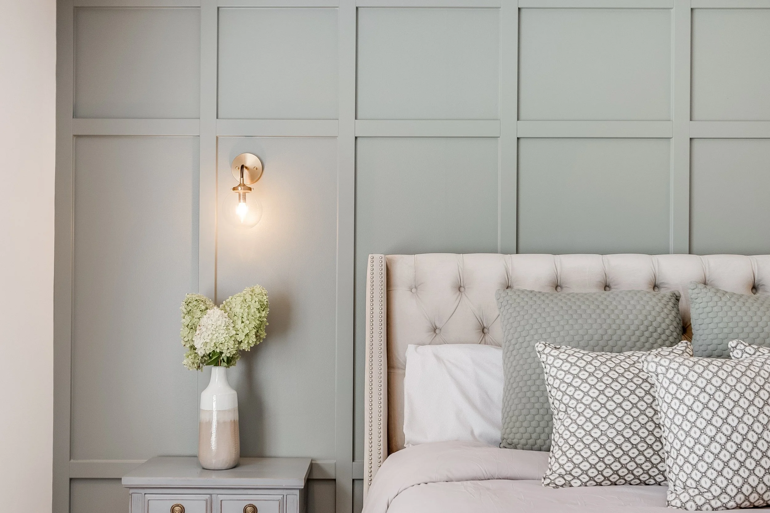 A bedroom with a light green paneled wall, a white tufted headboard, and six decorative pillows in shades of gray and white. A white bedside table holds a white vase with green hydrangeas, and a wall-mounted light with a bare bulb is above the vase.