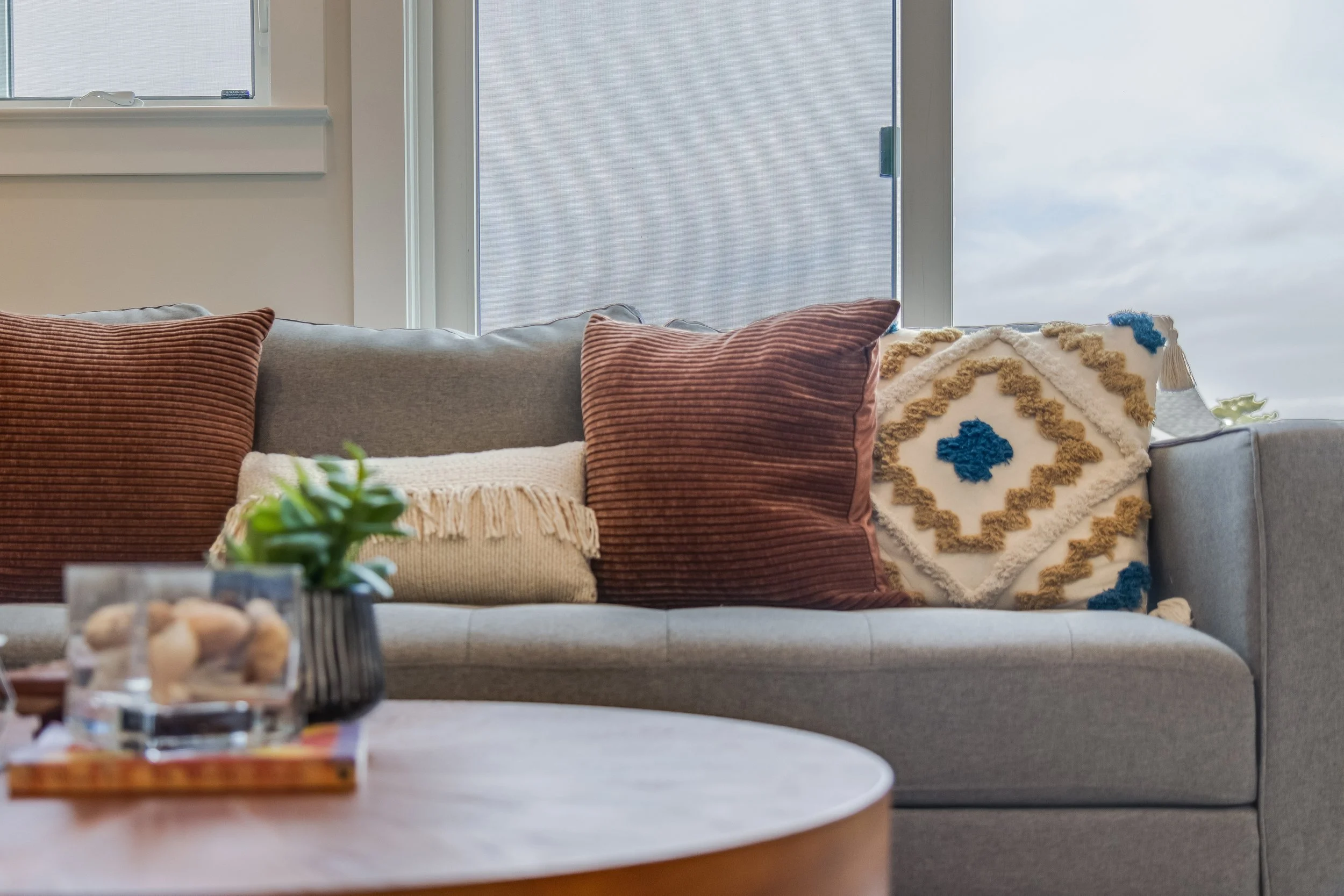A gray sofa with multiple decorative pillows in shades of brown, cream, and white with blue accents, in a living room with large windows.