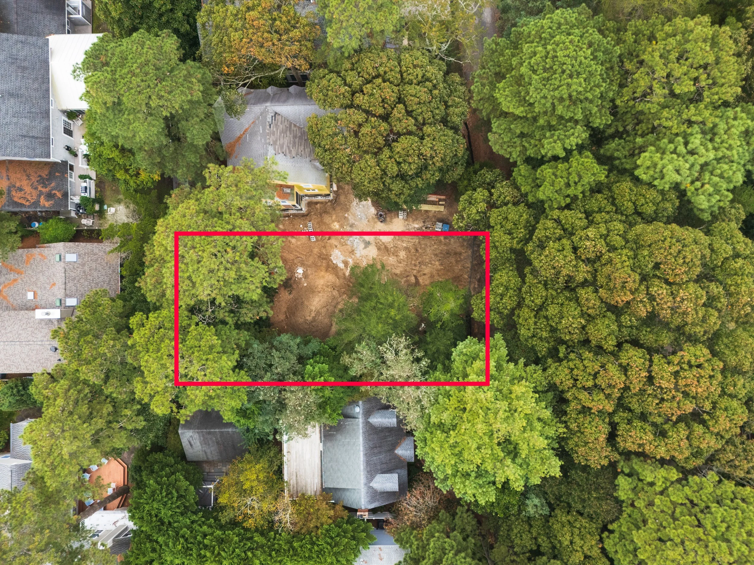 An aerial view of a residential neighborhood with a cleared rectangular plot of land outlined in red, surrounded by trees and neighboring houses.