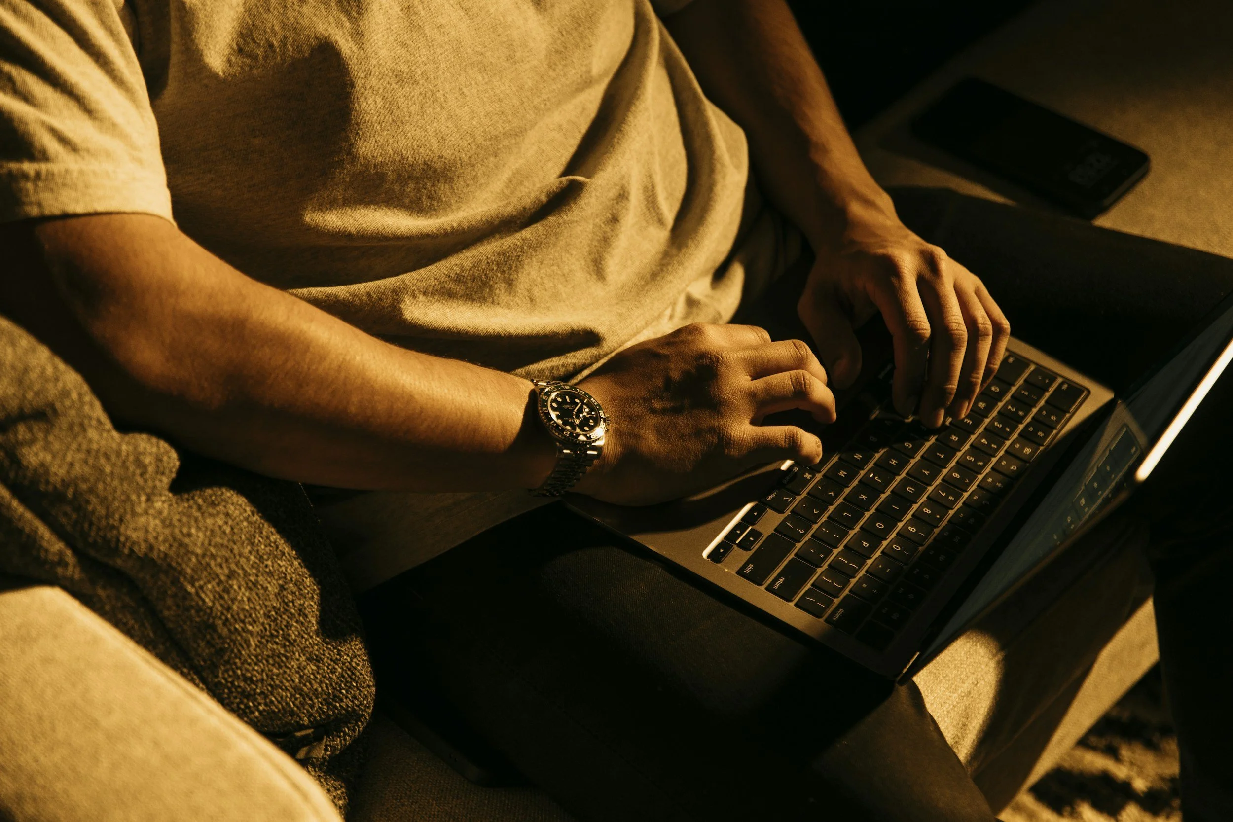 A person sitting on a couch using a laptop, wearing a beige T-shirt and a wristwatch, with a smartphone nearby in a dimly lit room.