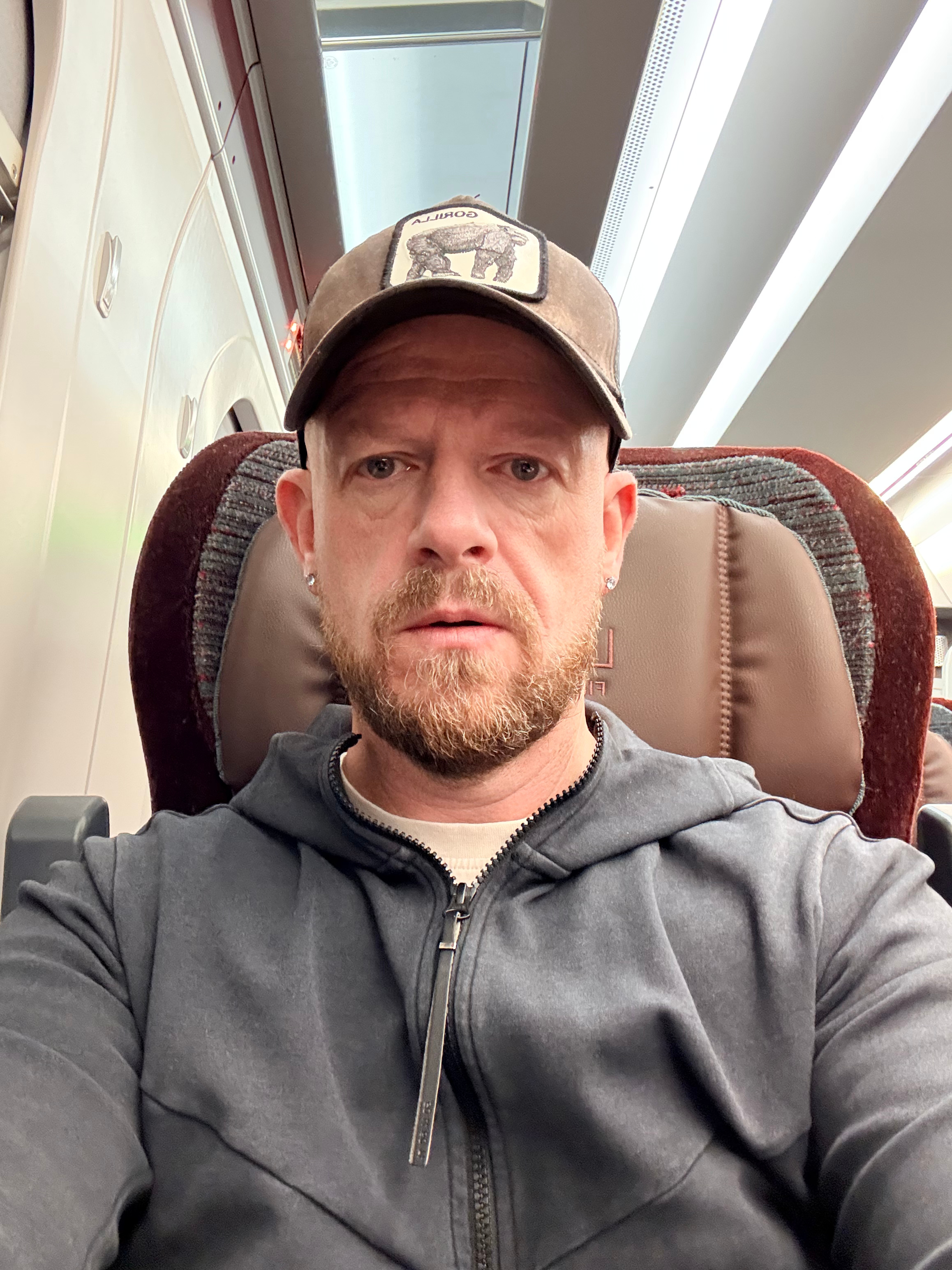 A man with a beard and earrings taking a selfie on an airplane, wearing a gray hoodie and a baseball cap with a bear patch.