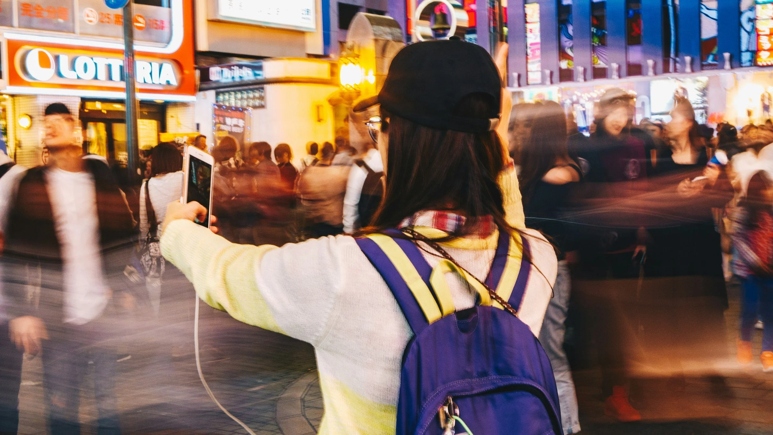 A woman taking a photo with her smartphone in a busy city street at night with many blurred pedestrians and bright neon signs.