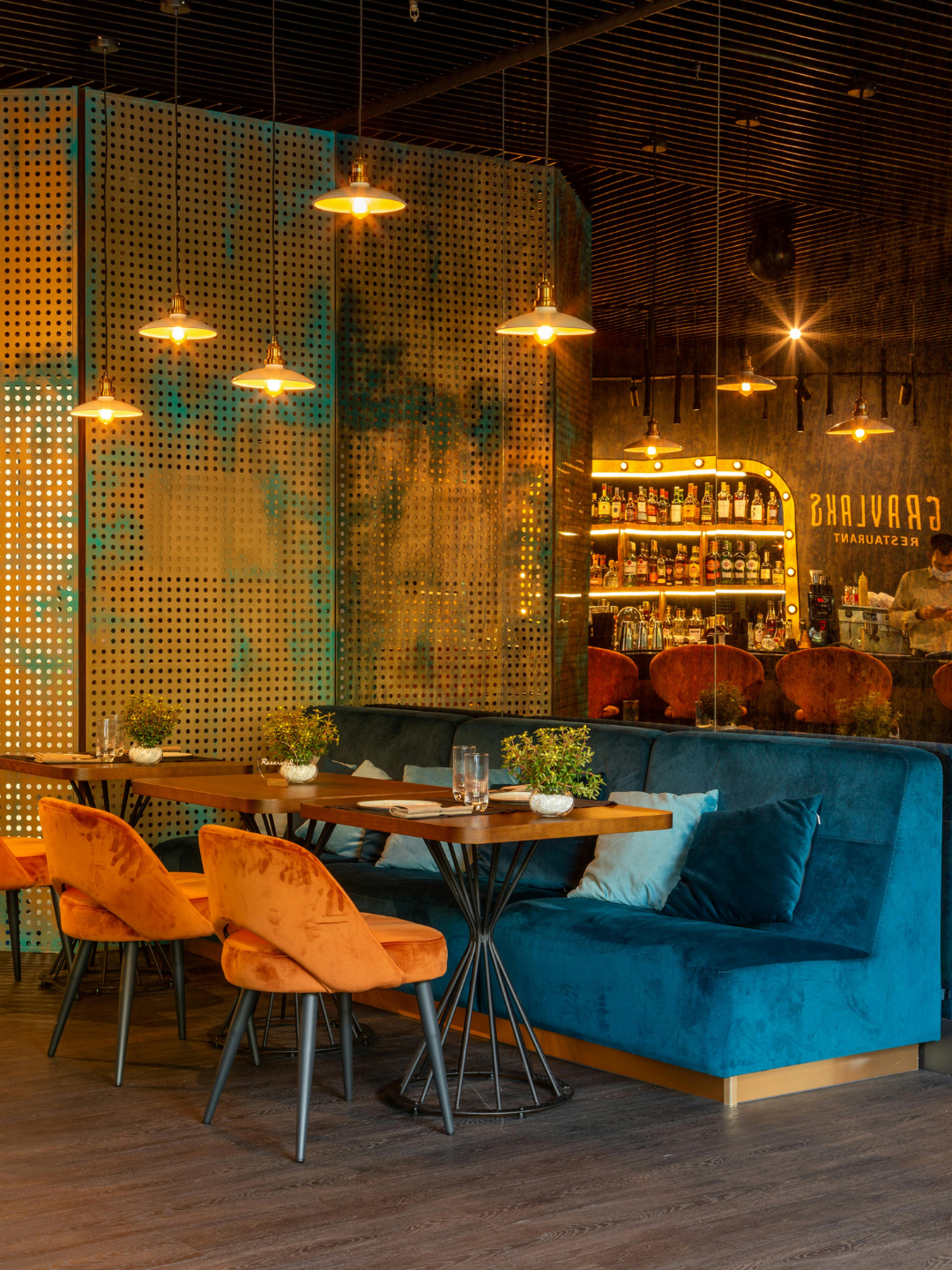 Interior view of a stylish bar and restaurant with a blue velvet seating area, wooden tables with small potted plants, orange upholstered chairs, and a stocked bar with illuminated shelves in the background. Hanging pendant lights provide warm lighting.