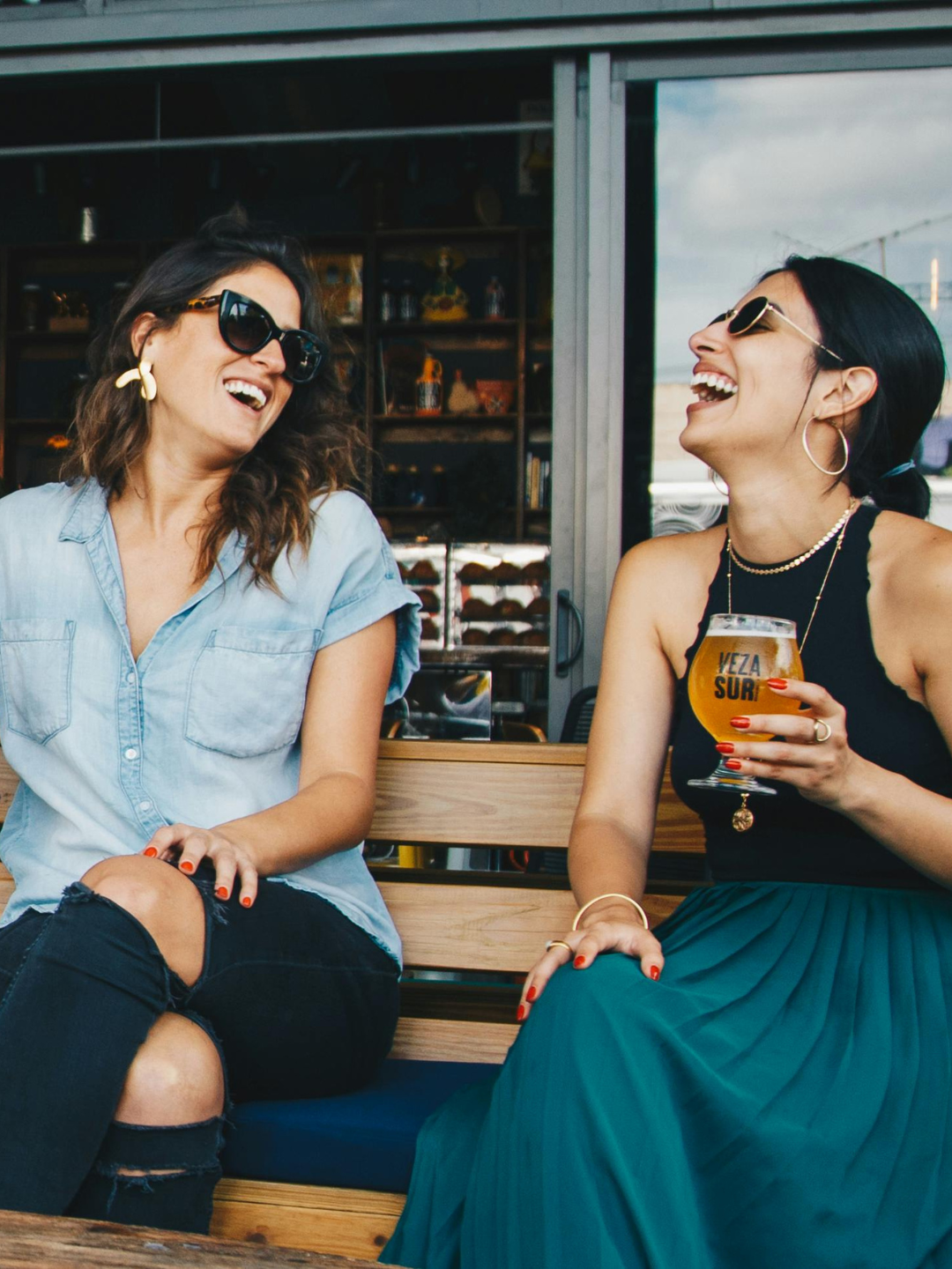Two women sitting on a wooden bench, laughing, with one holding a glass of beer. They are wearing sunglasses and casual stylish clothing.
