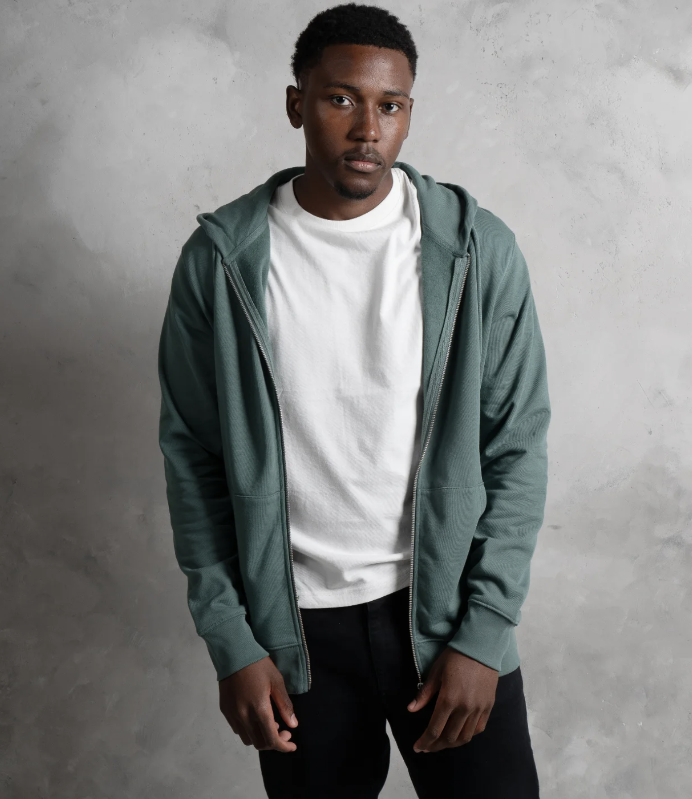 A young man with dark skin and short curly hair is standing against a light grey textured wall. Product photography setup inside The Space MCR studio in Manchester.