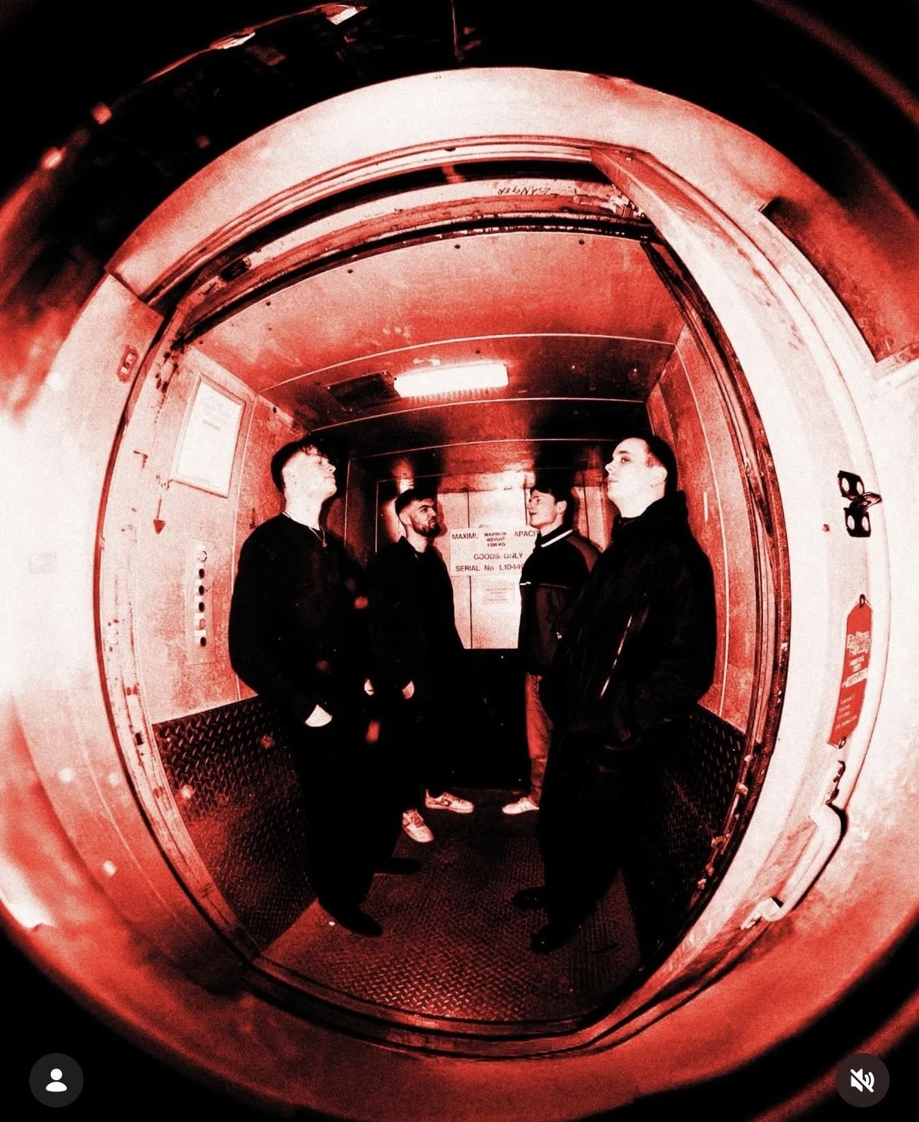 Four young men standing inside an elevator with red lighting, looking at each other and the walls around them.