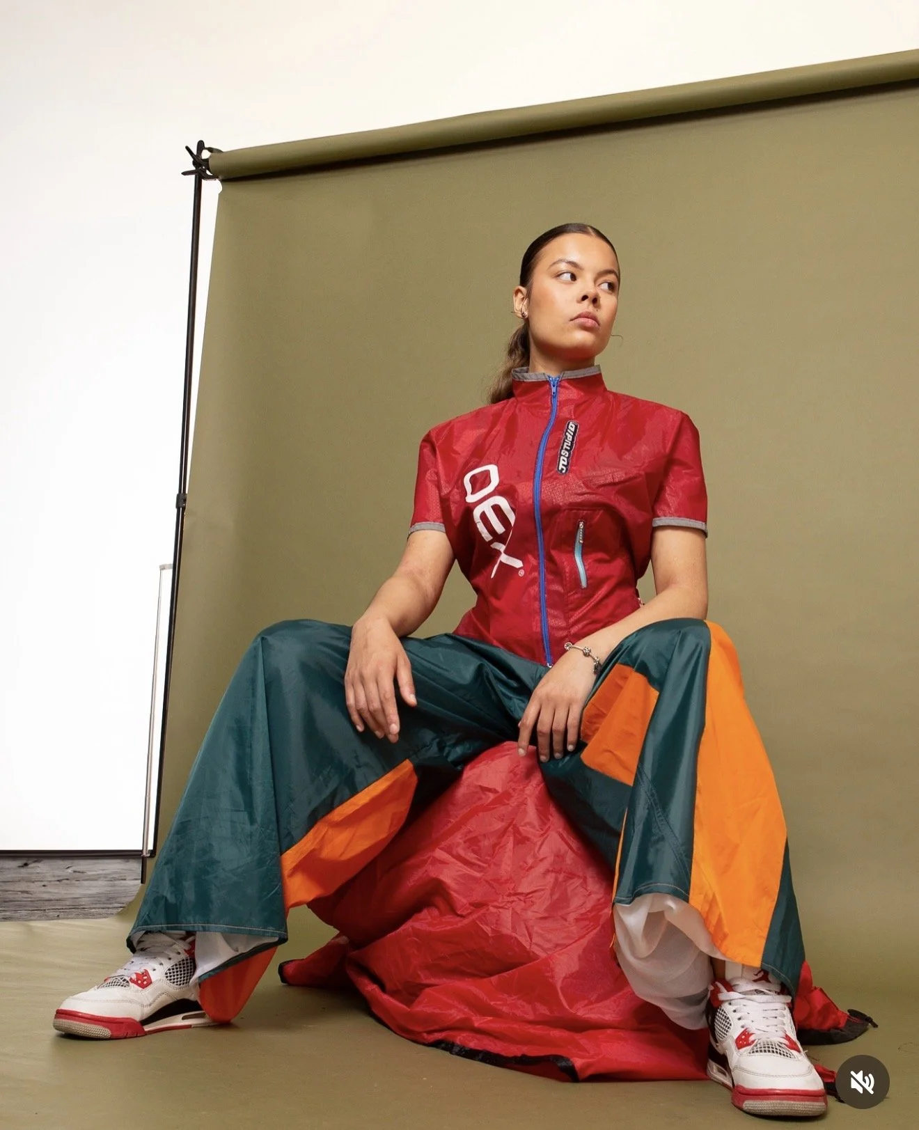 A woman sitting on a red bag in front of a green backdrop, wearing a colorful windbreaker, loose pants, and sneakers, looking to the side with a serious expression.