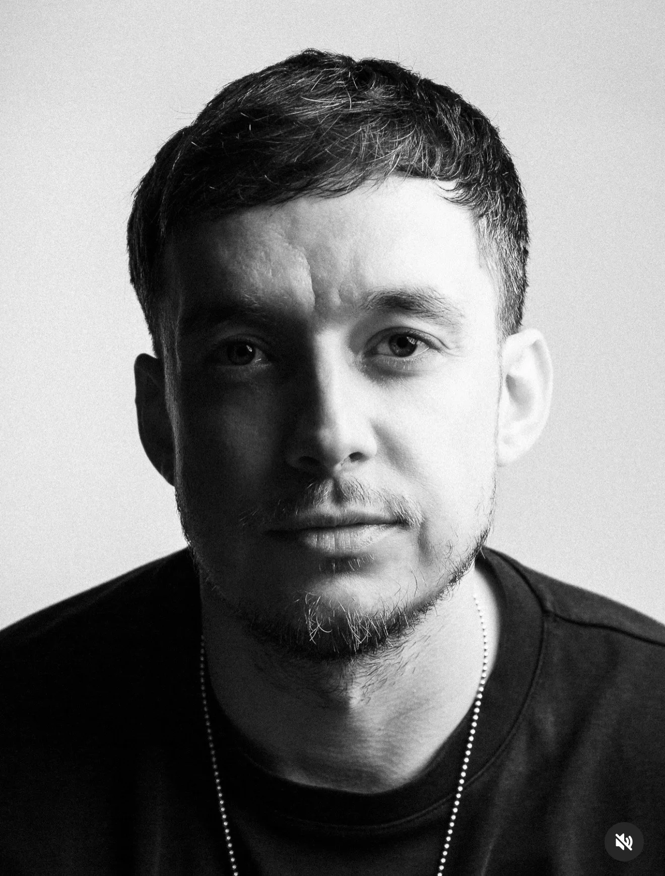 Black and white portrait of a young man with short hair and slight facial hair, wearing a dark shirt and a necklace, with a neutral expression.